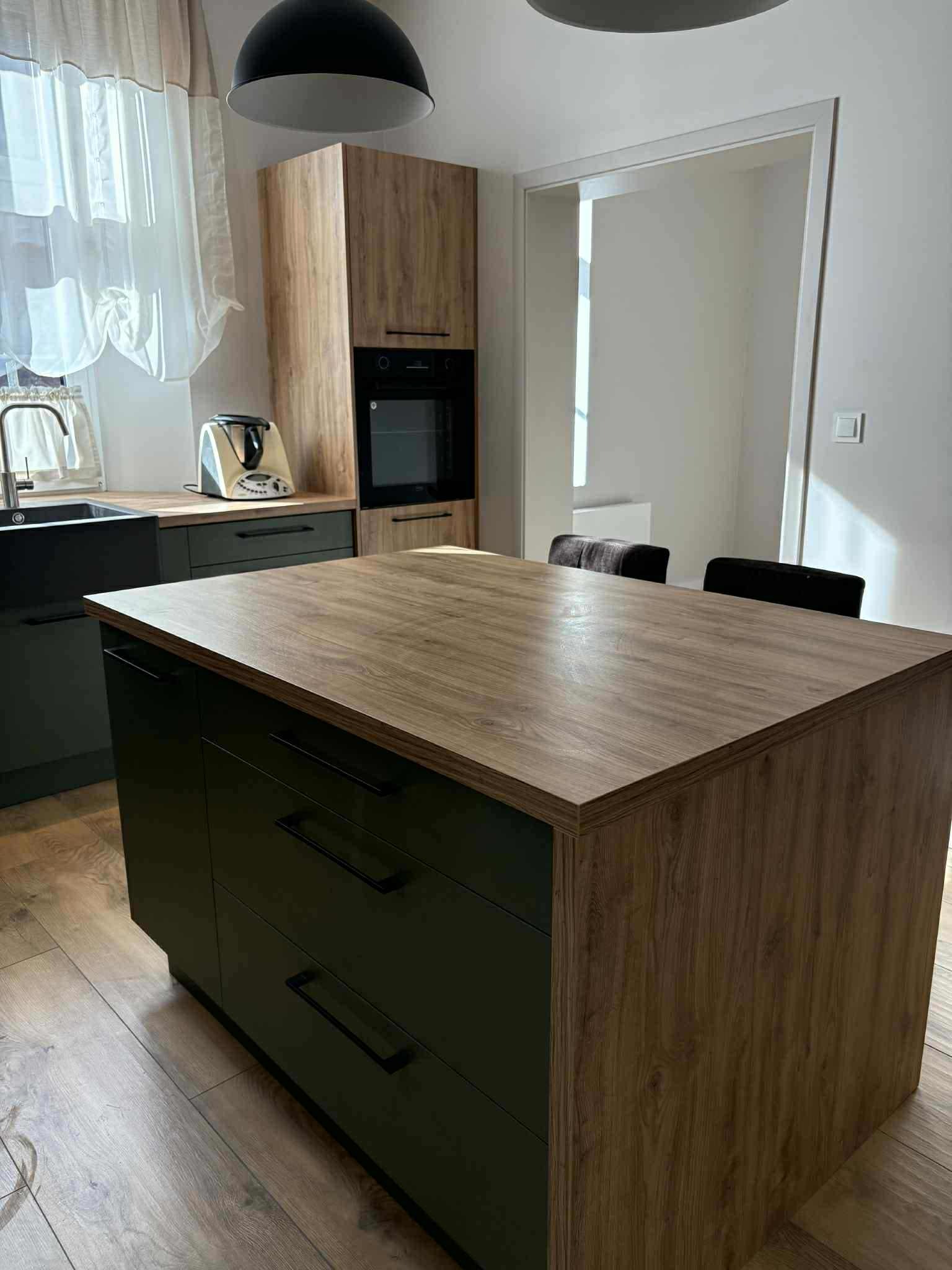 Nowoczesna wyspa kuchenna z drewnianym blatem i ciemnozielonymi szafkami, w tle zabudowa kuchenna z piekarnikiem i ekspres do kawy na blacie.