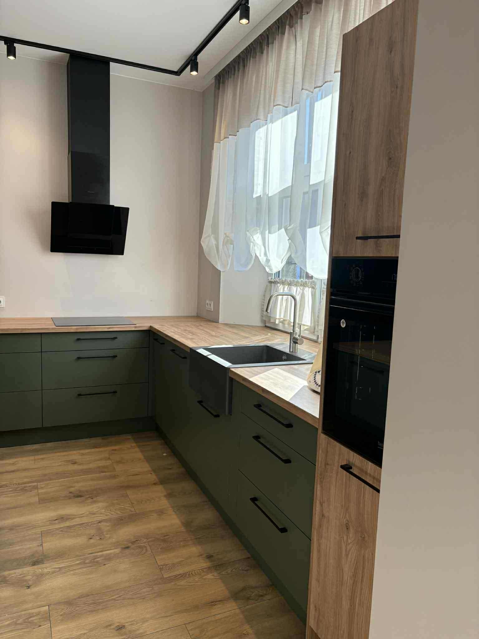 Nowoczesna kuchnia z zielonymi szafkami, drewnianym blatem i czarnym okapem. Ujęcie z perspektywy, ukazujące fragment okna z firanką.