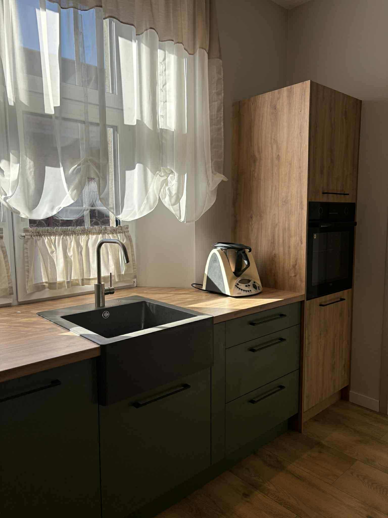 Nowoczesna kuchnia z zielonymi szafkami, drewnianym blatem i piekarnikiem w zabudowie. Okno z firankami w tle. Na blacie stoi robot kuchenny.