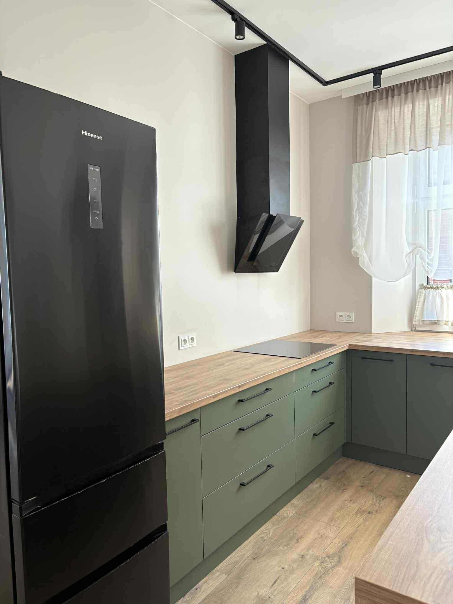 Nowoczesna kuchnia z zielonymi szafkami, drewnianym blatem i czarną lodówką Hisense. Minimalistyczny okap i oświetlenie punktowe dopełniają wystrój.