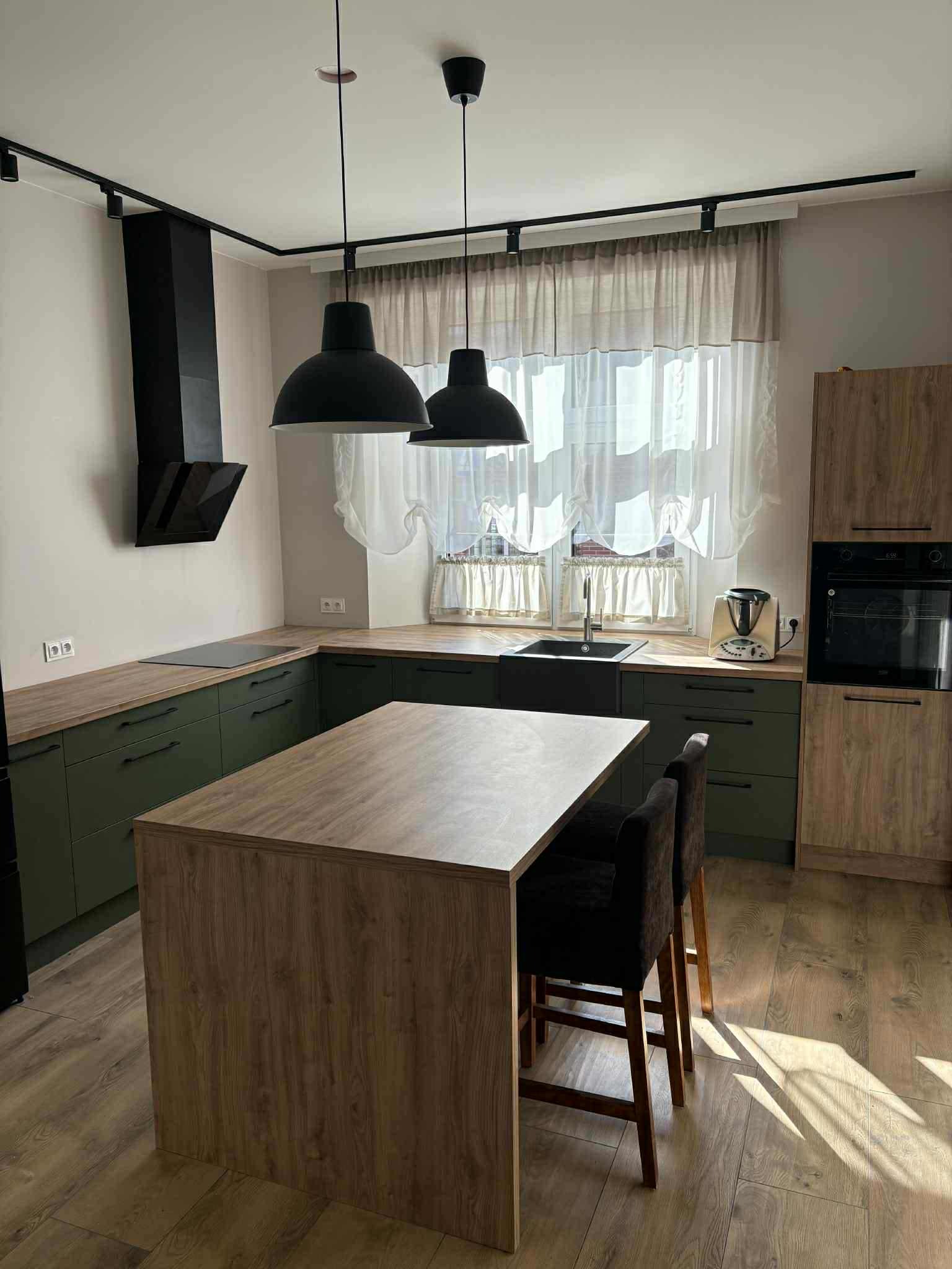 Nowoczesna kuchnia z wyspą, zielonymi szafkami i drewnianymi akcentami, oświetlona lampami wiszącymi. Widok na okno z firankami i sprzęt AGD.