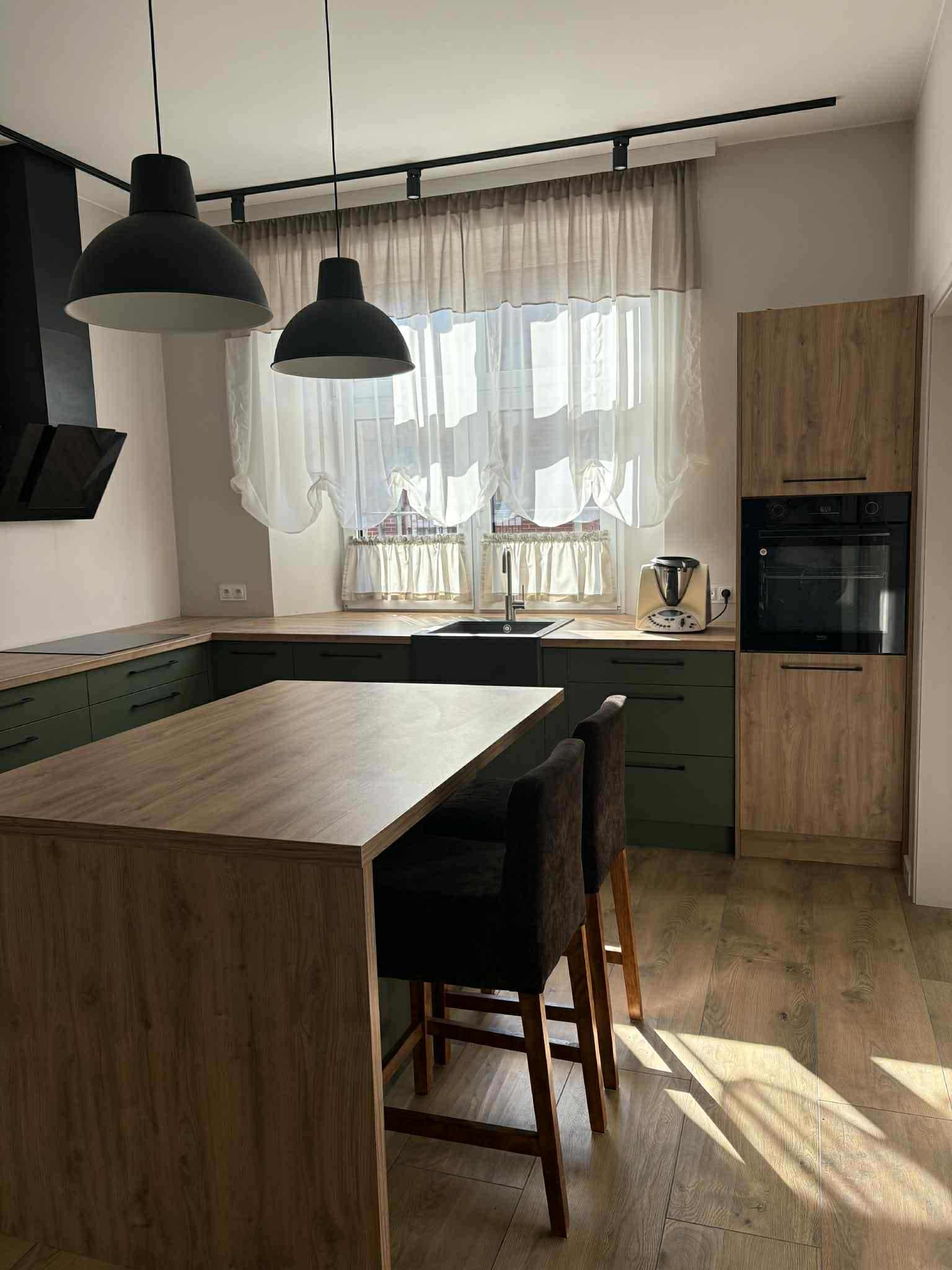 Nowoczesna kuchnia z wyspą, zielonymi szafkami i drewnianymi blatami. Nad wyspą wiszą dwie czarne lampy. Uwagę zwraca jasna podłoga i światło słoneczne.