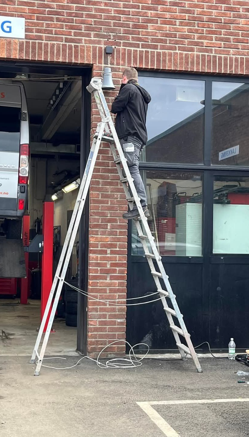 Mężczyzna na aluminiowej drabinie ustawia szarą lampę zewnętrzną na ceglanym budynku, widoczny kabel zasilający leżący na ziemi.