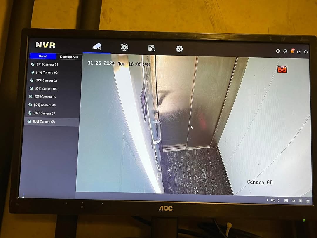 Ekran monitora AOC wyświetlający obraz z kamery monitoringu wewnątrz windy, zarejestrowany 25 listopada 2024 o godzinie 16:05:48, z widocznym interfejsem oprogramowania NVR i listą kamer.