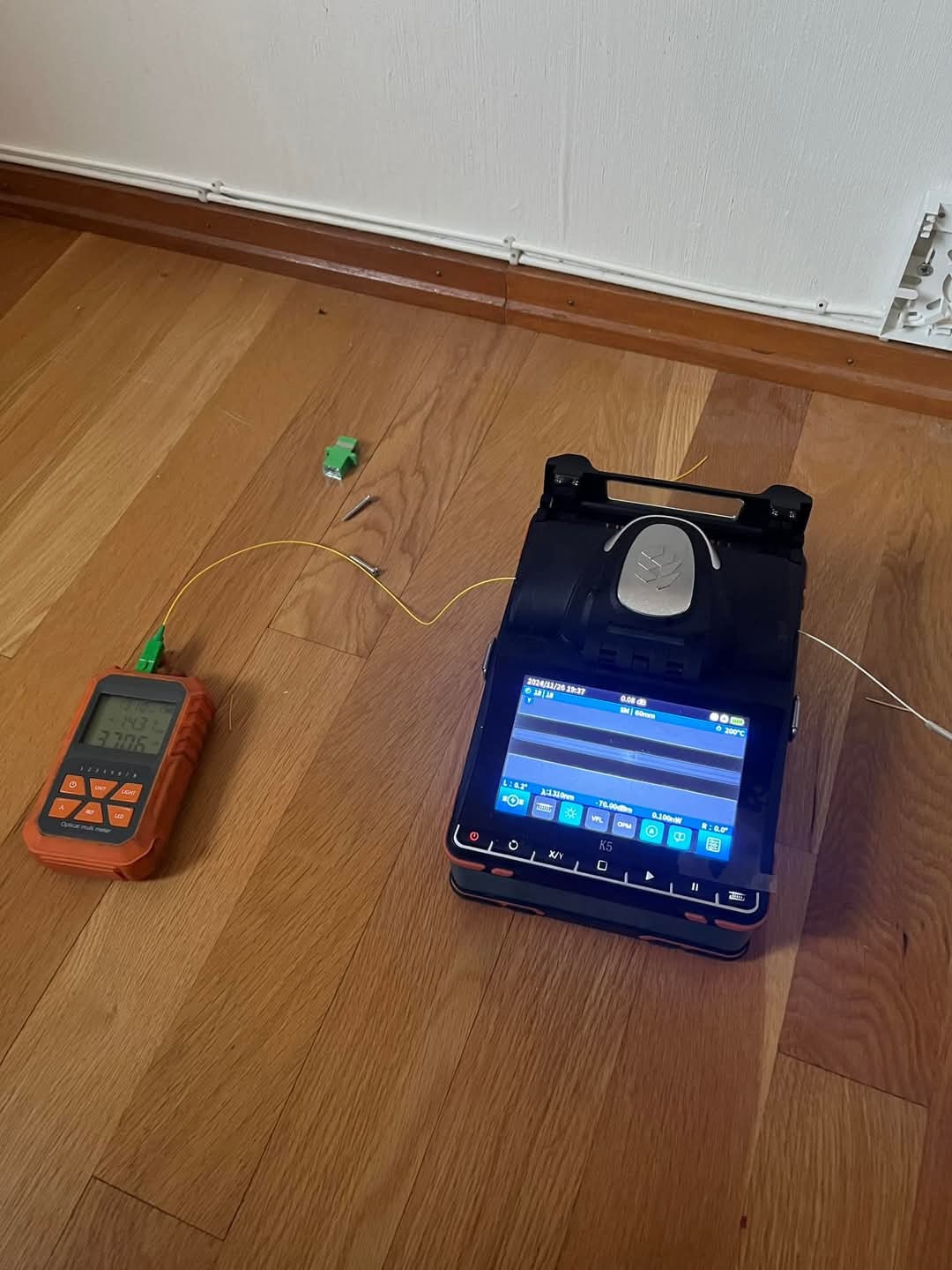 Urządzenie do spawania światłowodów z podłączonym miernikiem mocy optycznej na drewnianej podłodze, widoczne drobne elementy montażowe i fragment kabla światłowodowego.