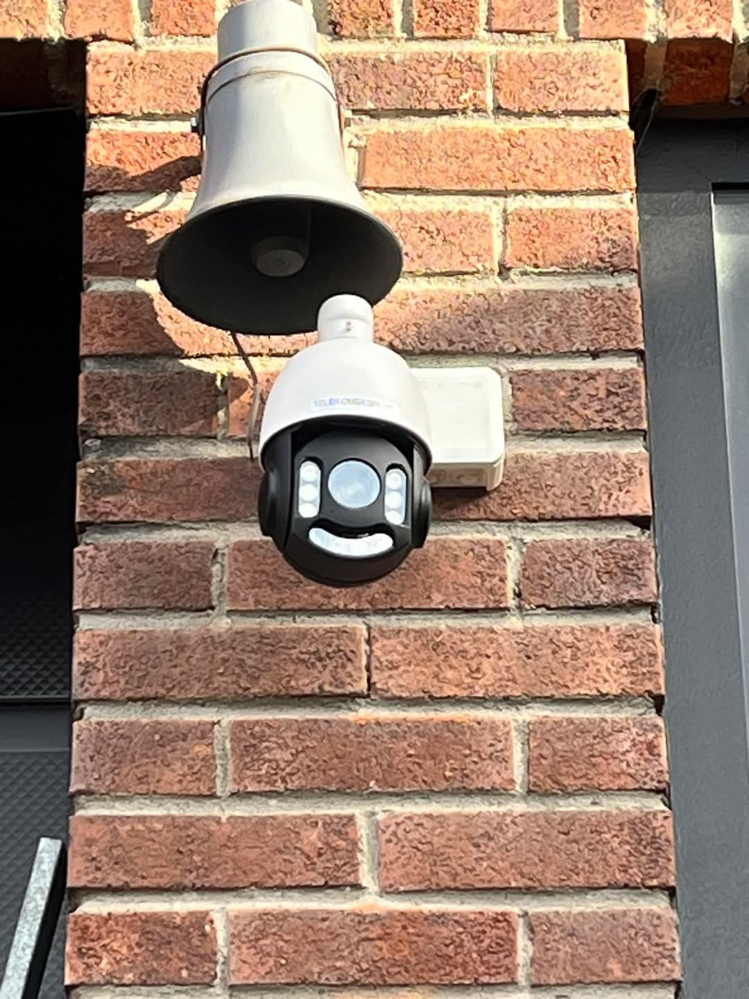 Zewnętrzna kamera obrotowa z głośnikiem alarmowym zamontowana na ceglanej ścianie budynku. Kamera w kolorze czarnym z białymi elementami. Widoczny fragment metalowej rury.
