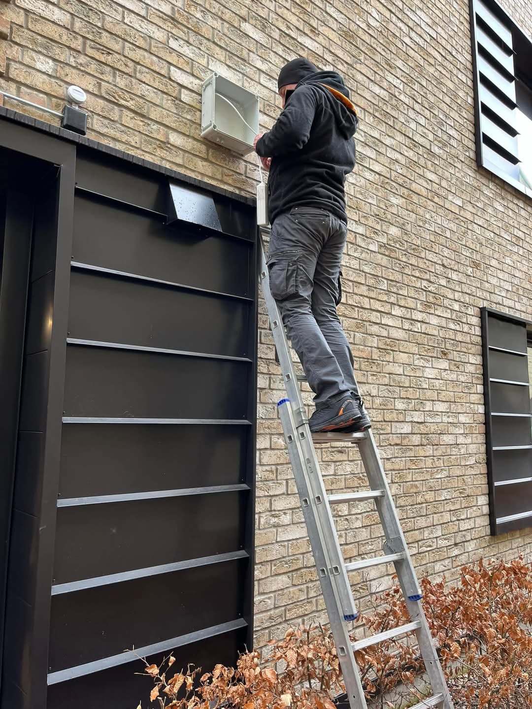 Elektryk na drabinie instaluje białą puszkę z kablami na ceglanej ścianie budynku z czarnymi żaluzjami okiennymi.