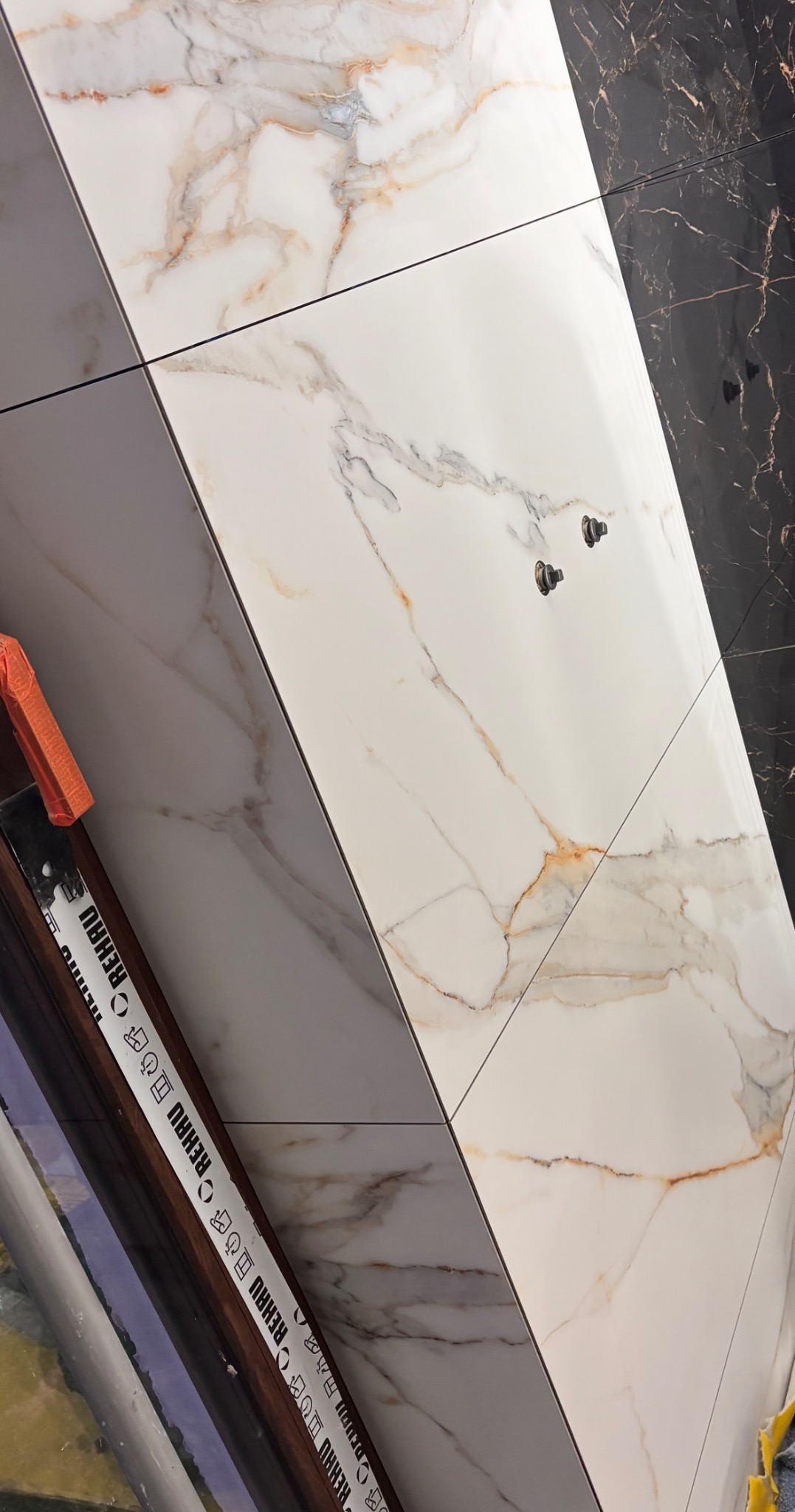 Ukończona ściana z płytek imitujących marmur w łazience, z widocznymi elementami instalacji wodnej i częściowo widoczną taśmą ochronną REHAU.