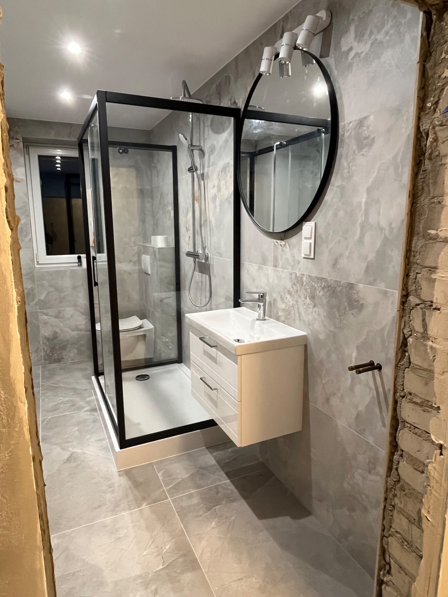 Wykończona łazienka w szarościach z kabiną prysznicową z czarnymi ramami, owalnym lustrem z oświetleniem i białą szafką pod umywalką, widoczne fragmenty surowej ściany z cegły.