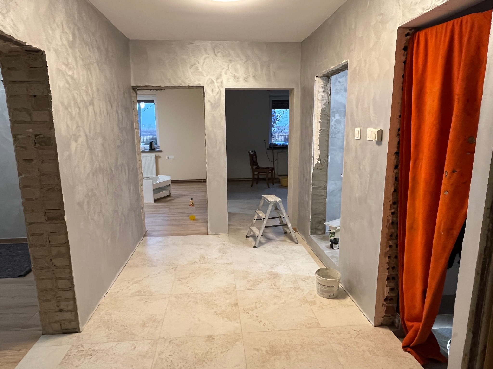 Korytarz w trakcie prac wykończeniowych: ściany pokryte dekoracyjną, srebrzystą strukturą, widoczne ślady po kuciu ościeżnic, pomarańczowa zasłona w przejściu, drabina aluminiowa i wiadro...