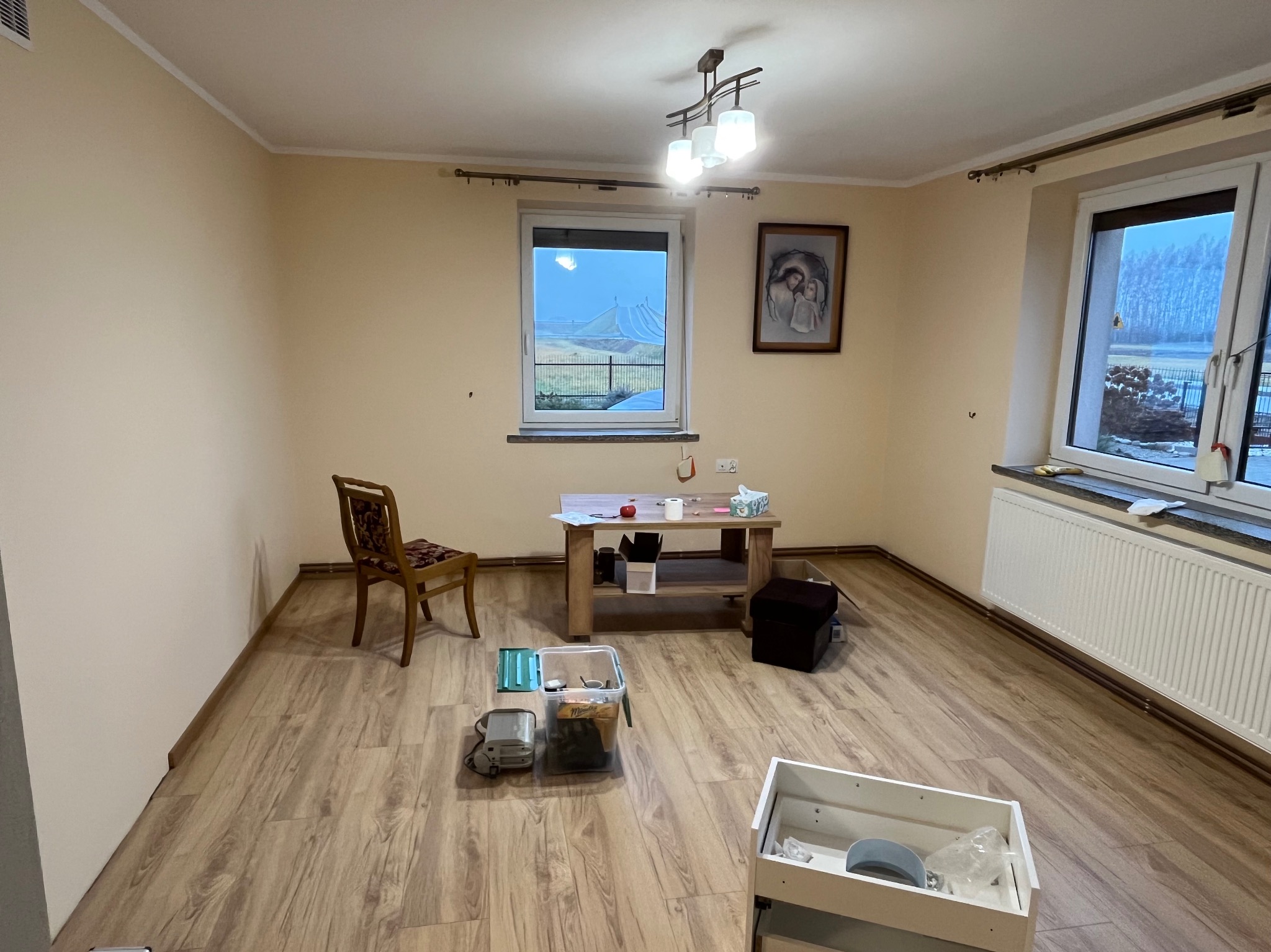 Pokój przed remontem: puste ściany w odcieniach beżu, drewniana podłoga, okna z widokiem na zimowy krajobraz, meble i narzędzia przygotowane do prac wykończeniowych, obraz na ścianie.