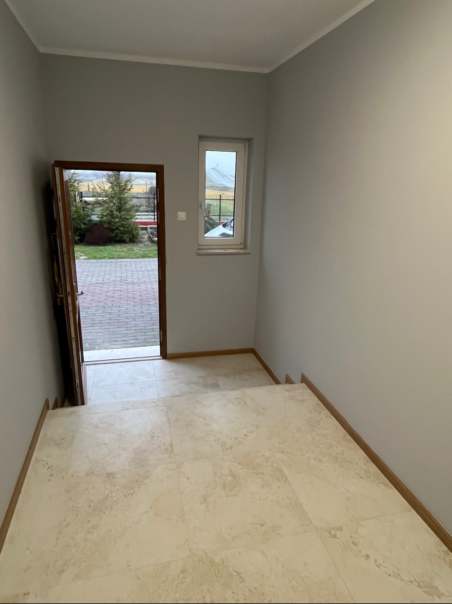 Jasne płytki podłogowe na schodach wewnątrz domu, świeżo pomalowane szare ściany, widok na zewnątrz przez otwarte drzwi i małe okno.