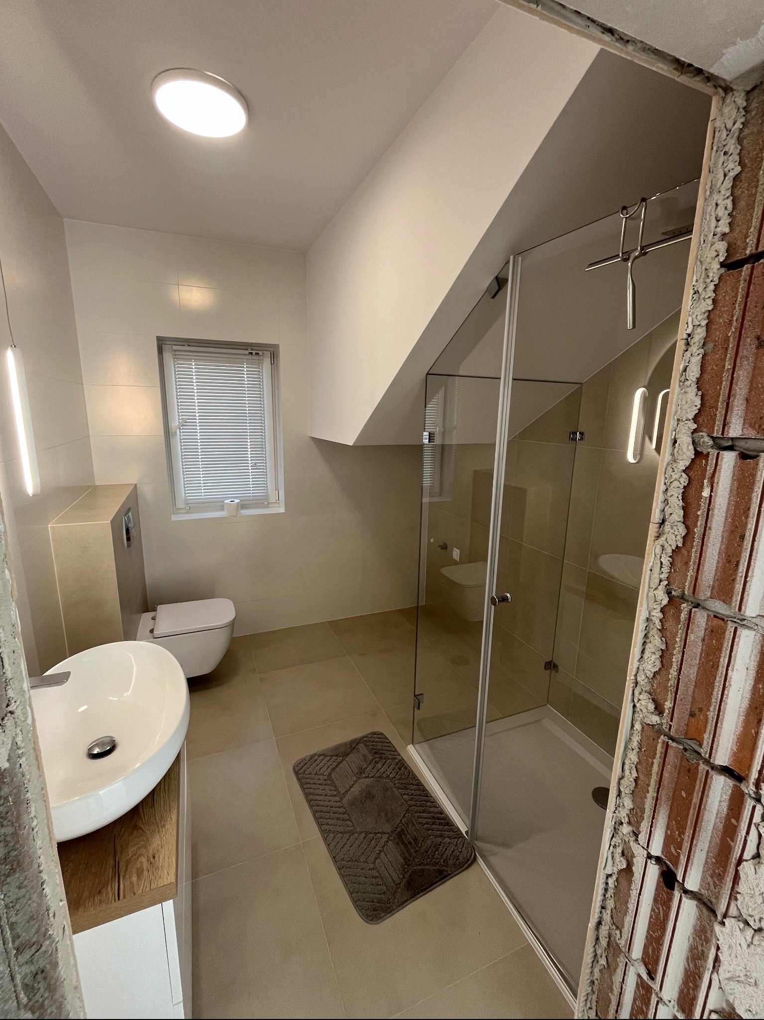 Nowoczesna łazienka z kabiną prysznicową ze szklanymi drzwiami, umywalką nablatową na drewnianej szafce, podwieszaną toaletą i oświetleniem LED, widok na surową ścianę z cegły po lewej stronie.