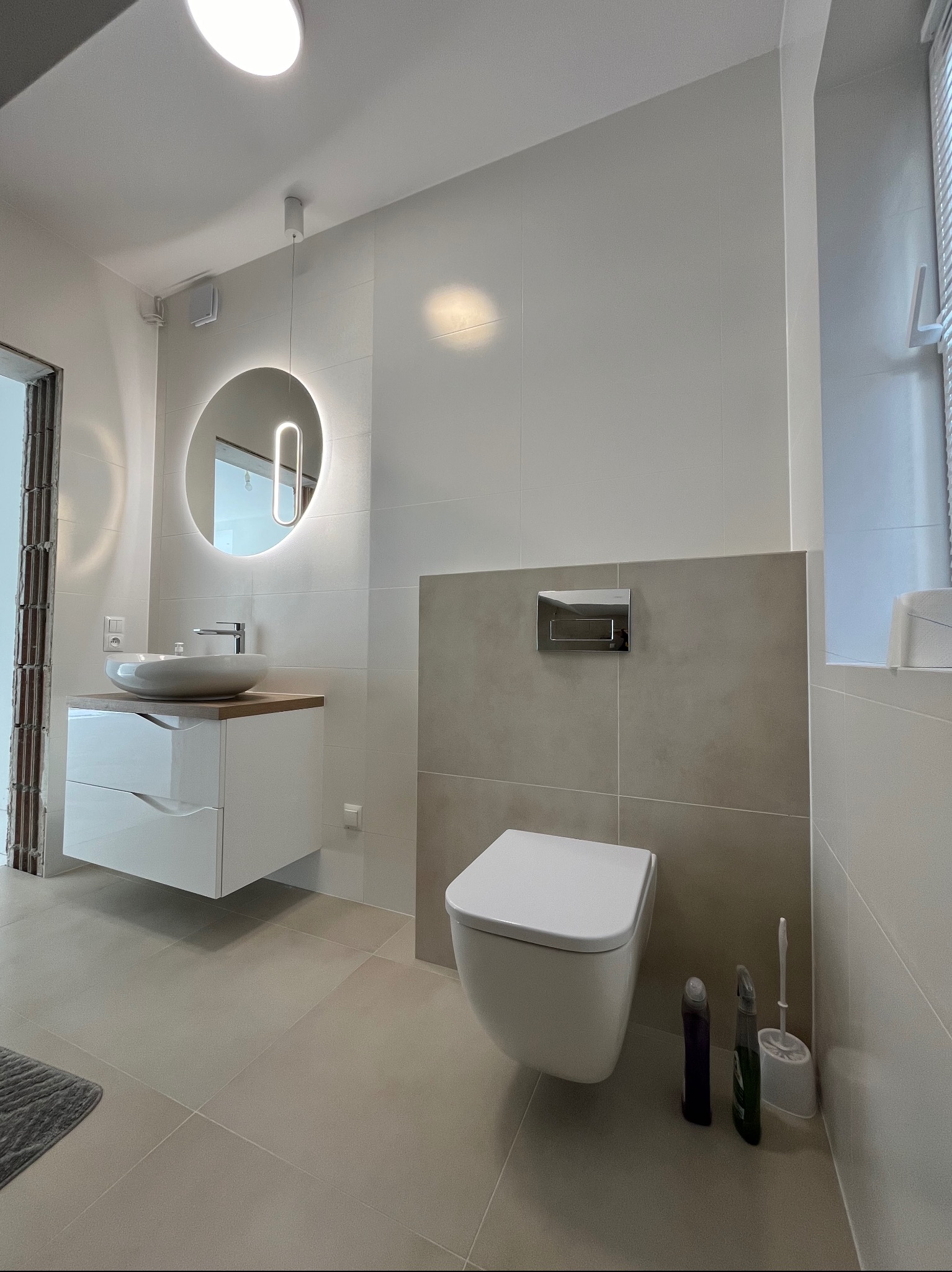 Nowoczesna łazienka z podwieszaną toaletą, umywalką nablatową z szafką i okrągłym lustrem z podświetleniem LED, wykończona jasnymi płytkami ceramicznymi.