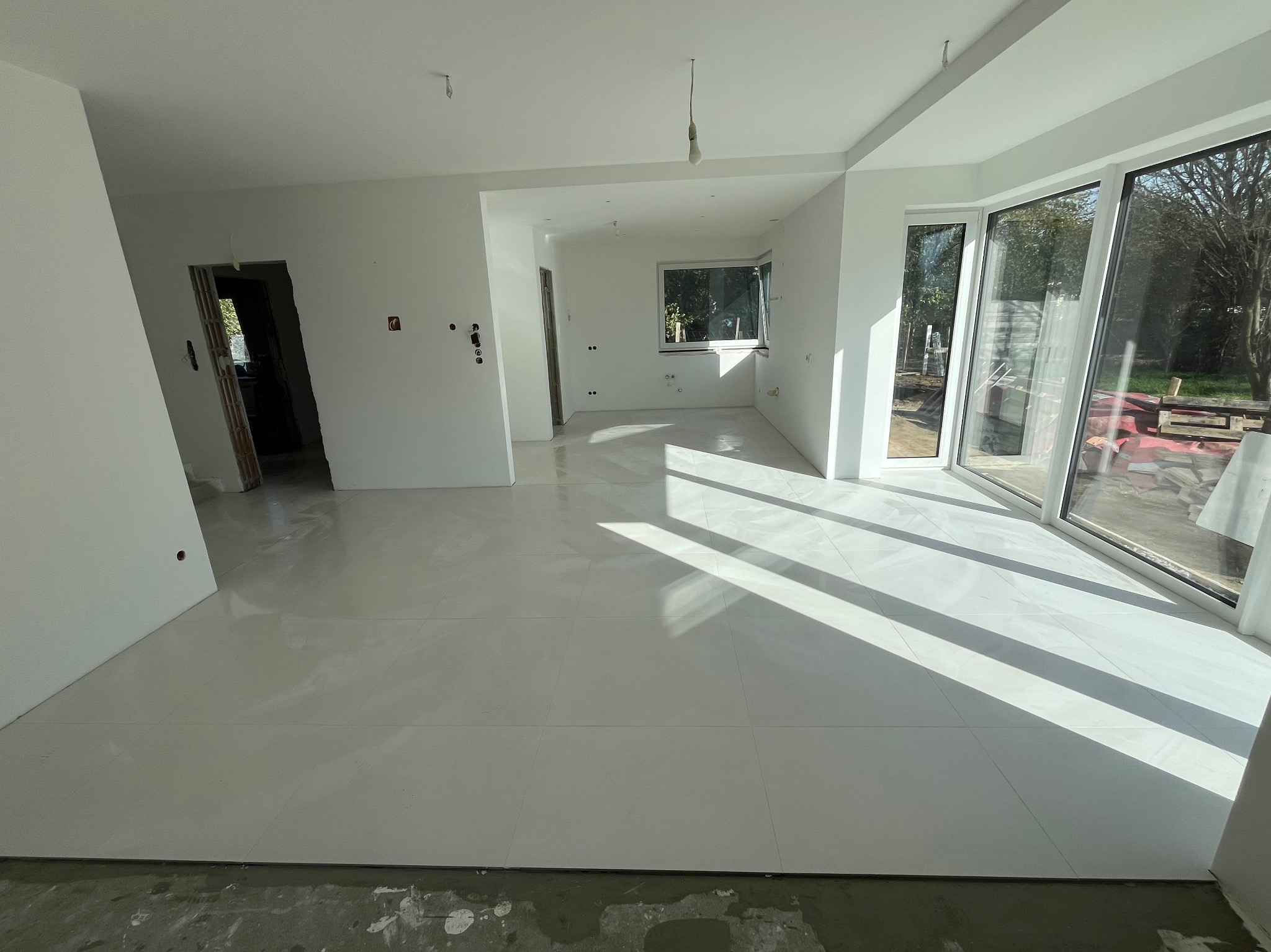 Jasne wnętrze domu w trakcie wykończenia, z nową białą podłogą z płytek, świeżo pomalowanymi ścianami i dużymi oknami wpuszczającymi silne promienie słoneczne.