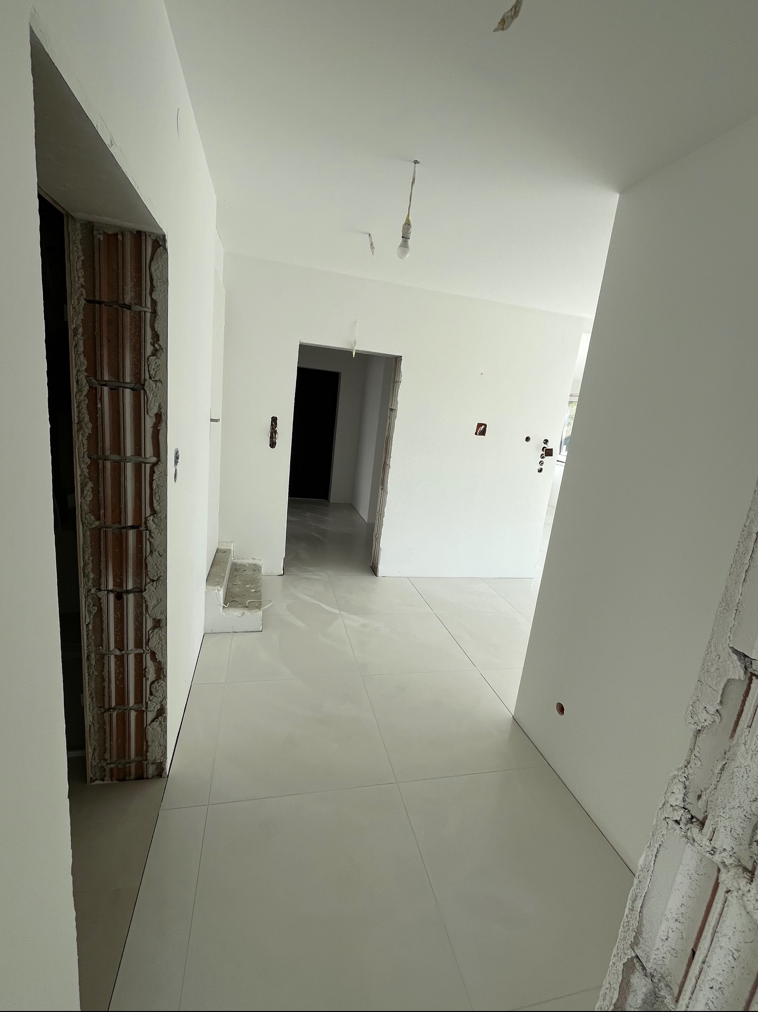 Wnętrze w trakcie remontu: białe ściany, jasna podłoga z płytek, niedokończone ościeżnice, widoczne instalacje elektryczne.