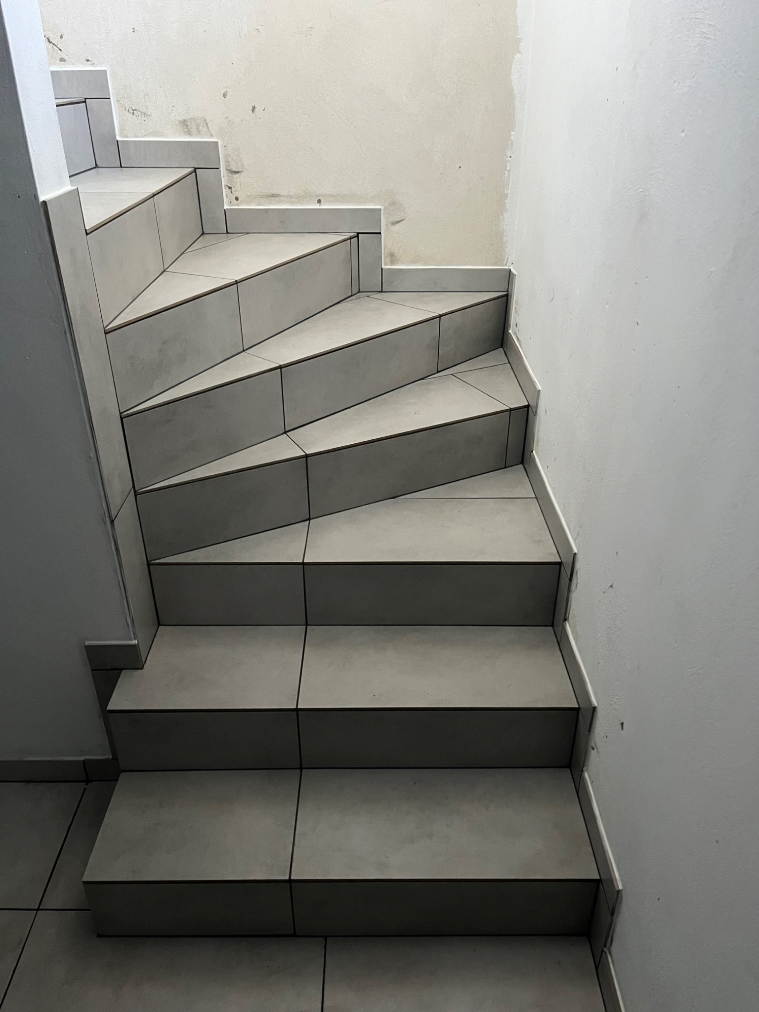 Wykończone schody z szarych płytek ceramicznych, widoczne fugi i niedokończone malowanie ściany w tle.