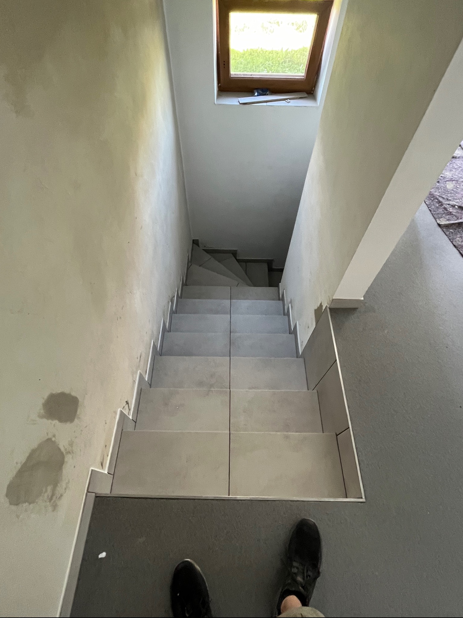Klatka schodowa w trakcie wykańczania: ułożone płytki na schodach z aluminiowymi listwami, surowe ściany z śladami po łataniu, okno dachowe w tle.