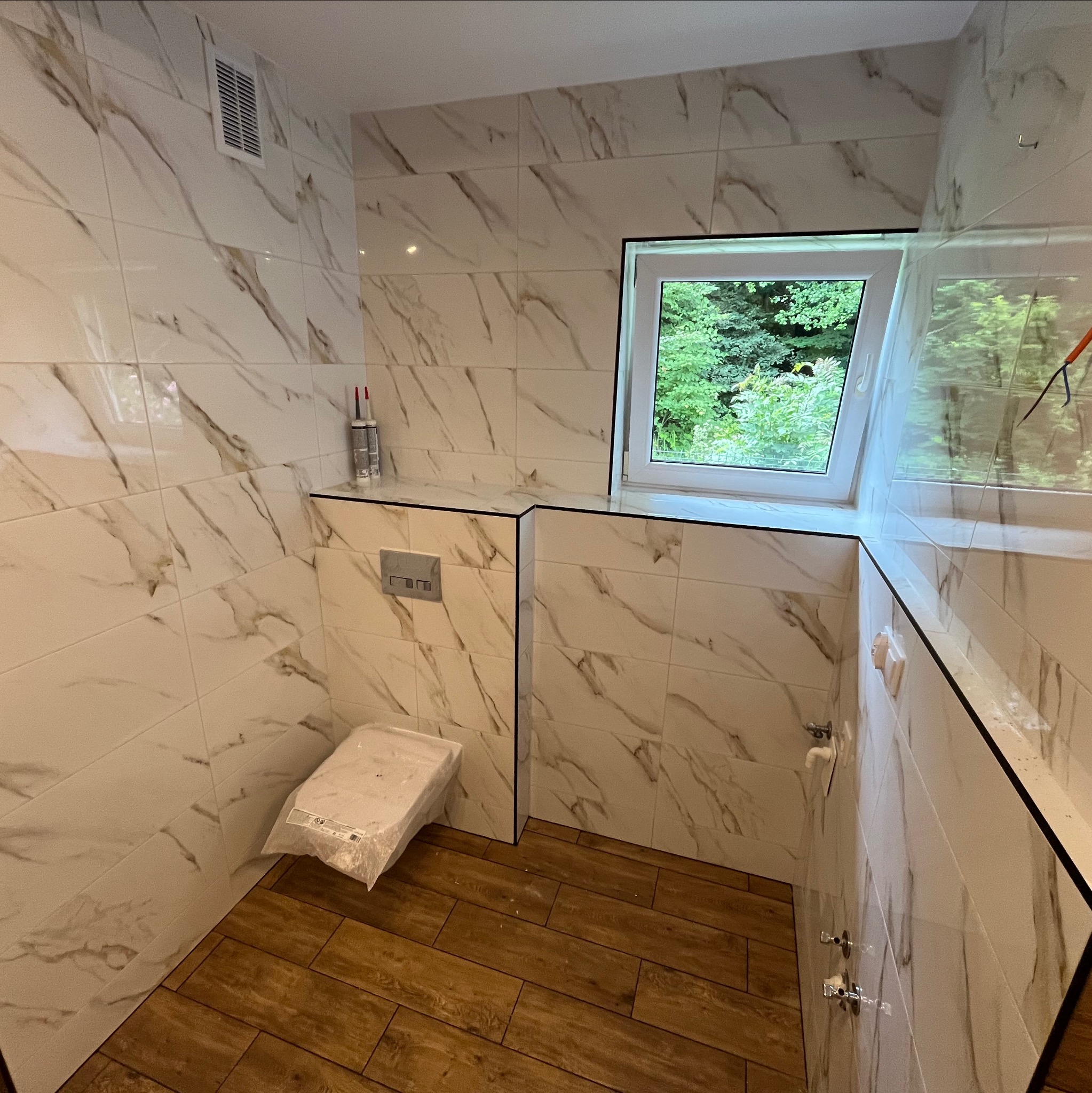 Wykończona łazienka z marmurowymi płytkami, drewnopodobną podłogą i oknem dachowym z widokiem na zieleń, widoczne elementy instalacji sanitarnej i częściowo zakryta toaleta.
