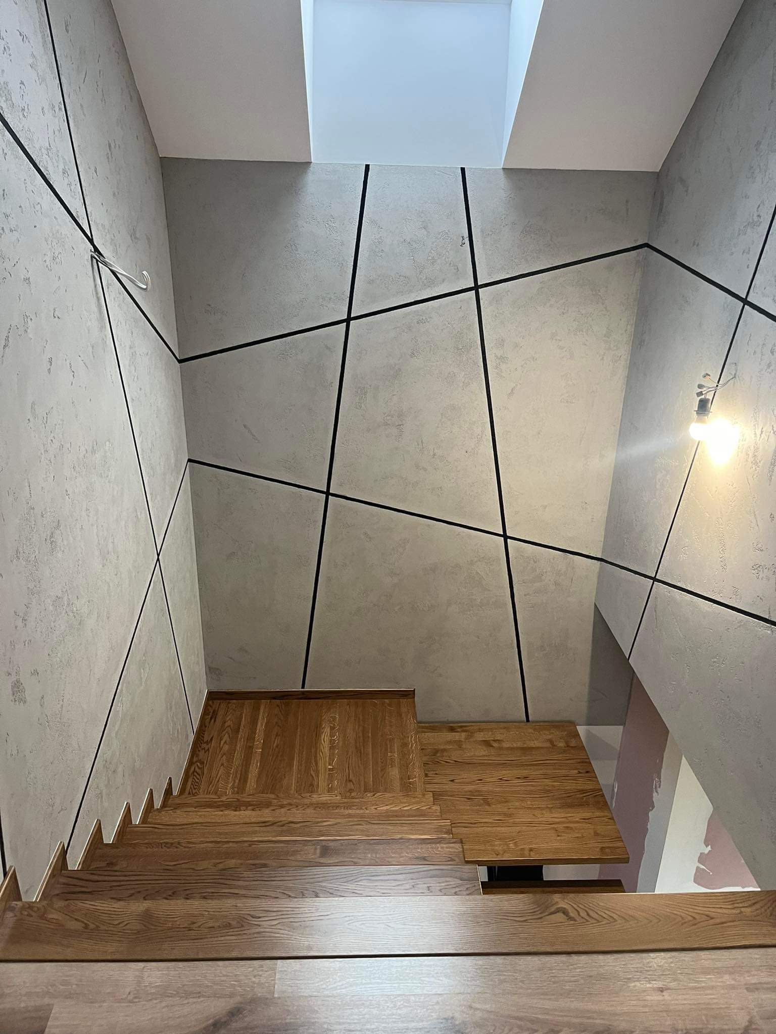 Klatka schodowa z drewnianymi schodami i szarymi ścianami ozdobionymi czarnymi liniami, widok z góry, oświetlenie naturalne z okna dachowego.