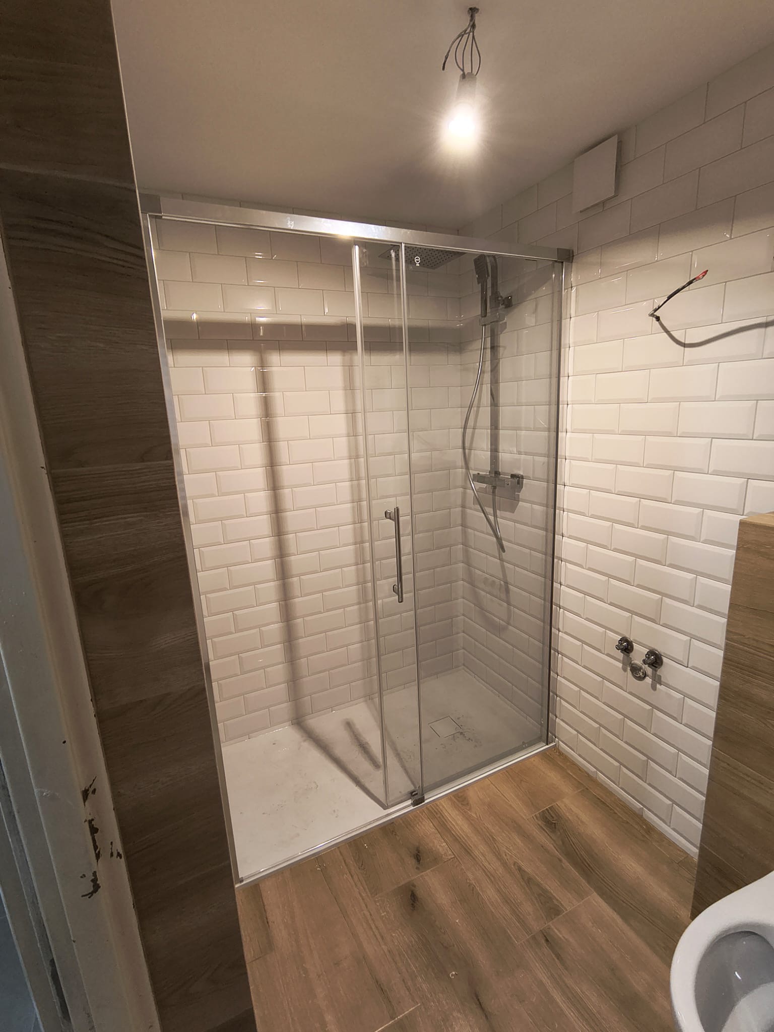 Nowoczesna kabina prysznicowa z przesuwnymi drzwiami, wykończona białymi, prostokątnymi płytkami i drewnopodobną podłogą. Widoczne elementy instalacji.