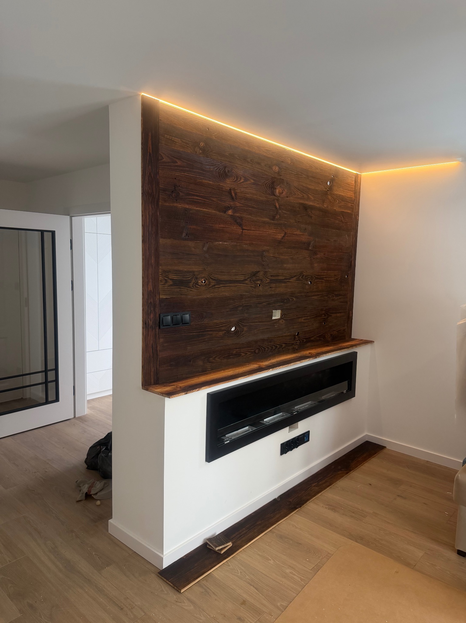 Drewniana ściana z oświetleniem LED i wbudowanym kominkiem elektrycznym w nowoczesnym wnętrzu. Widoczne gniazdka i włączniki. Podłoga wyłożona panelami.