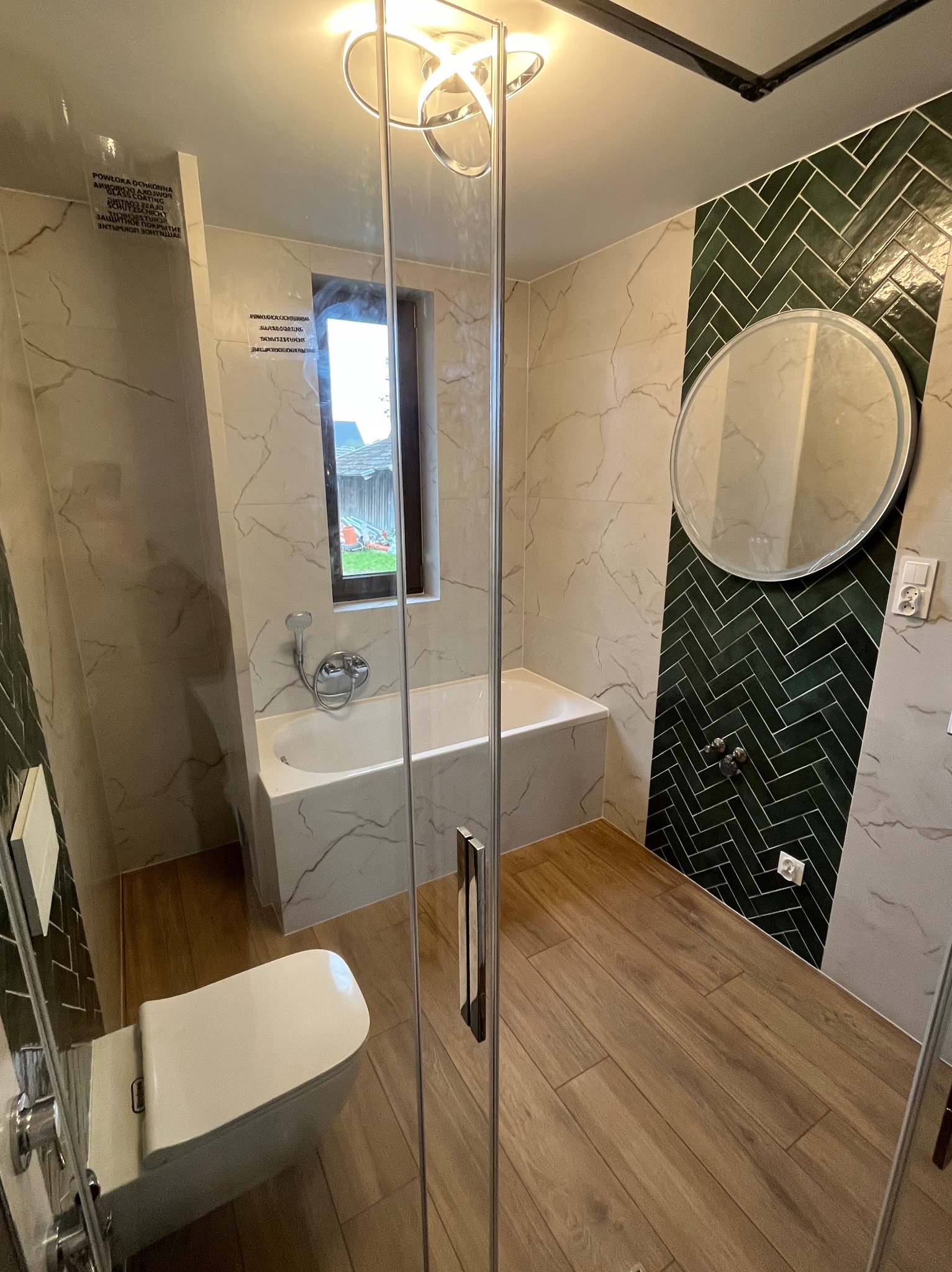 Nowoczesna łazienka z kabiną prysznicową, białą armaturą i zieloną ścianą w jodełkę. Jasne płytki imitujące marmur i drewnopodobna podłoga tworzą spójną całość.
