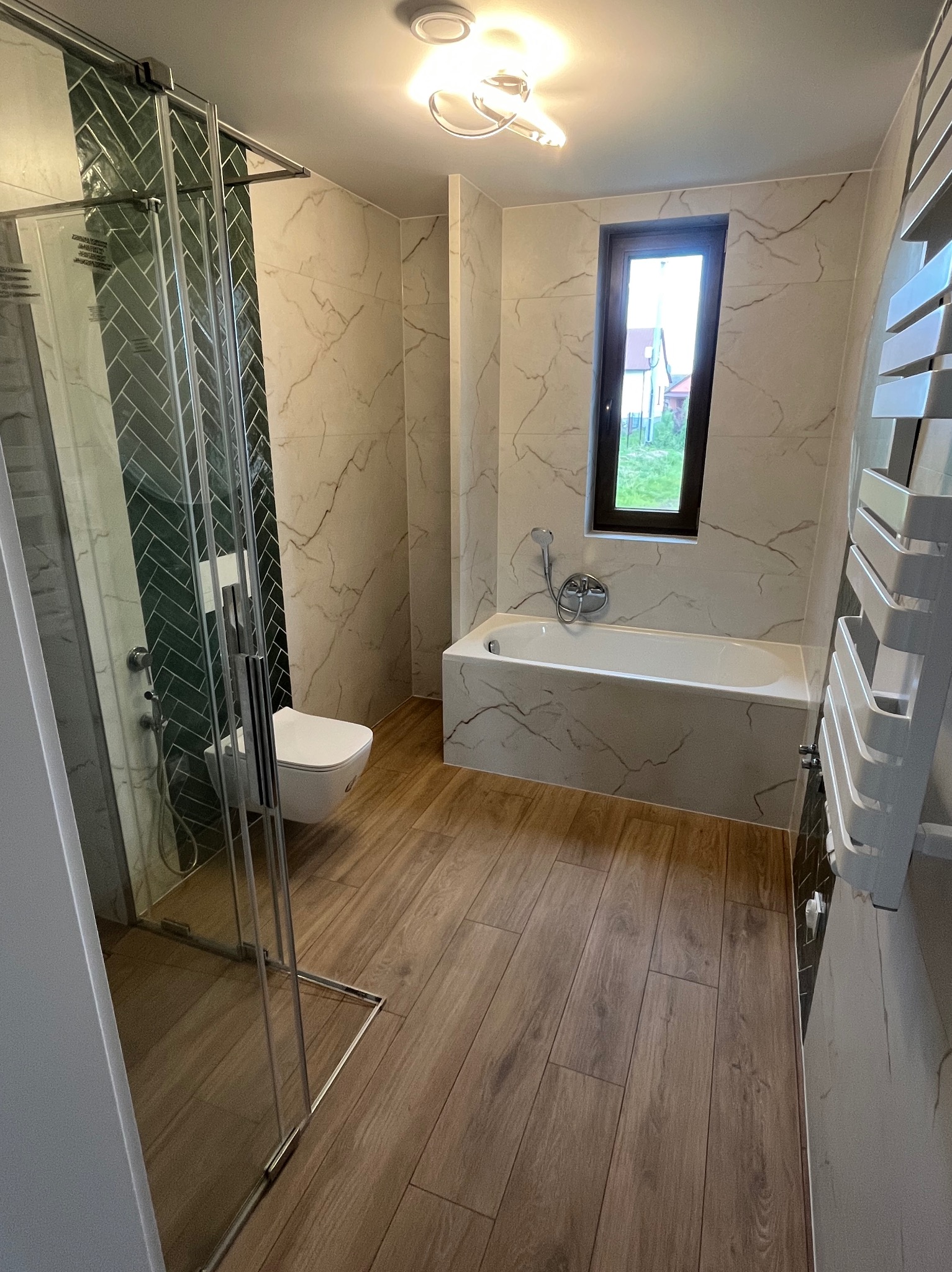 Nowoczesna łazienka z kabiną prysznicową, toaletą, wanną i grzejnikiem drabinkowym. Ściany wyłożone płytkami imitującymi marmur, podłoga drewnopodobna. W tle okno.