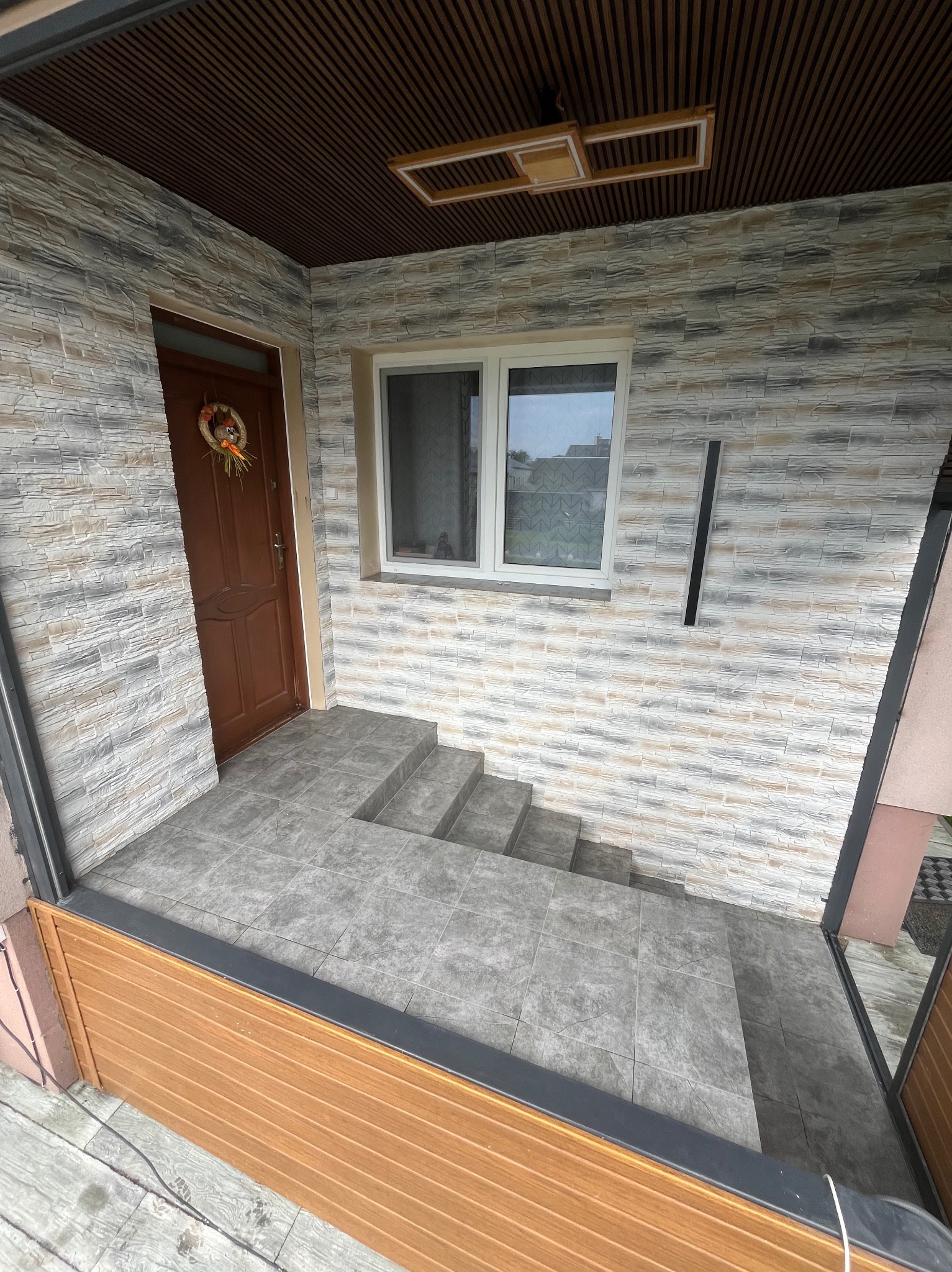 Wykończone wejście do domu z kamienną elewacją, brązowymi drzwiami z jesiennym wieńcem i szarymi płytkami na schodach. Widoczne okno i drewniany sufit.
