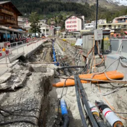 Otwarty wykop w miejskiej scenerii, ukazujący sieć rur i kabli w trakcie instalacji lub naprawy, z widocznymi elementami infrastruktury miejskiej w tle oraz górami w oddali.