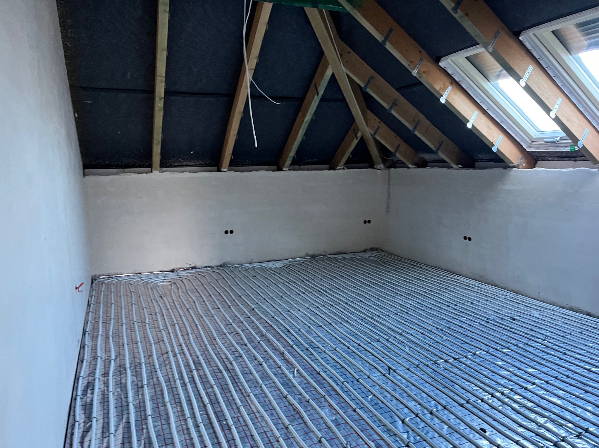 Równomiernie ułożone rury ogrzewania podłogowego na folii izolacyjnej, widoczne drewniane krokwie poddasza z oknem dachowym, surowe ściany w trakcie wykończenia.