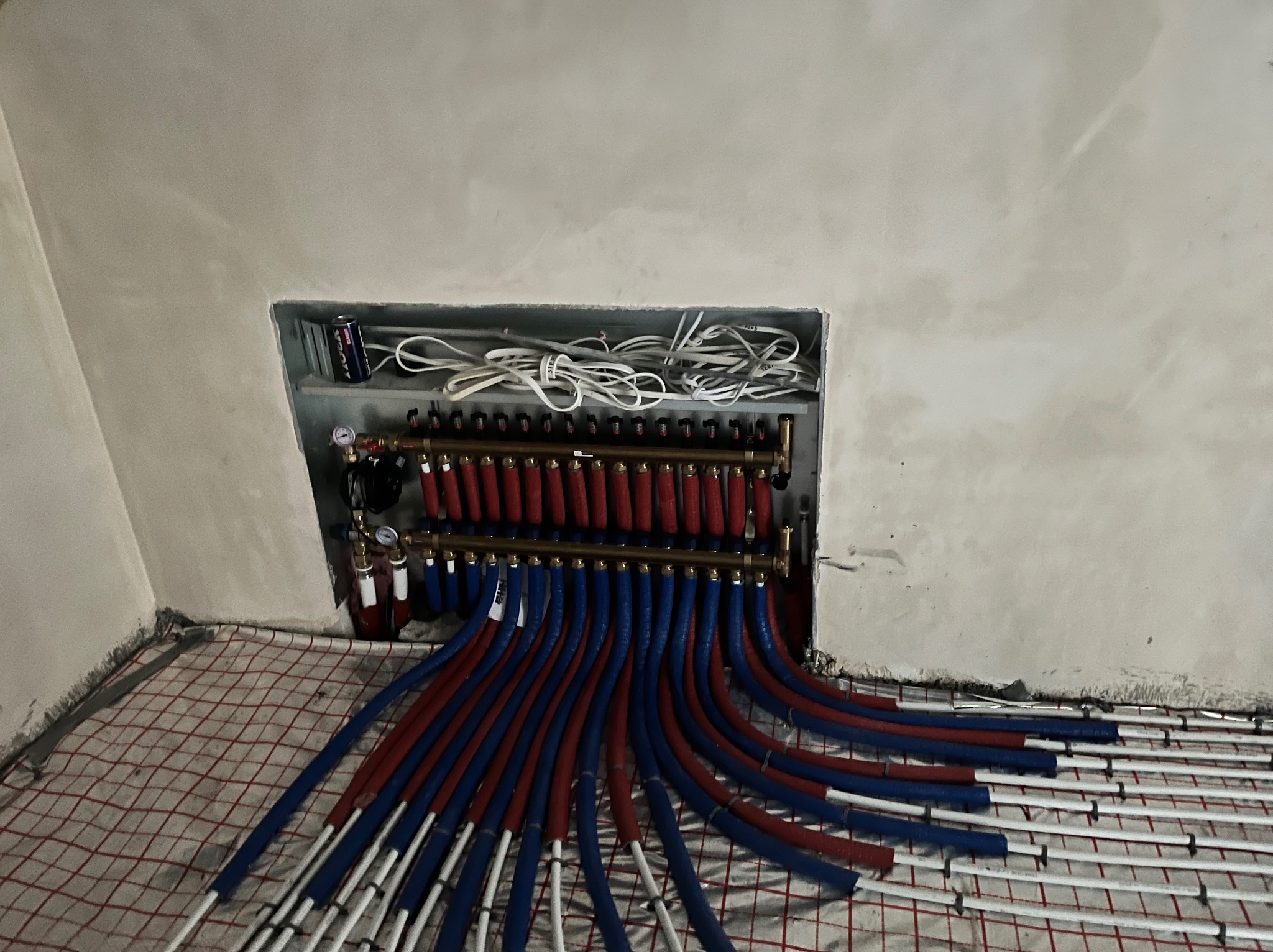 Rozdzielacz ogrzewania podłogowego z rurami w kolorach czerwonym i niebieskim, ułożonymi na siatce, widoczny wnęka w ścianie z przewodami elektrycznymi i puszką napoju.