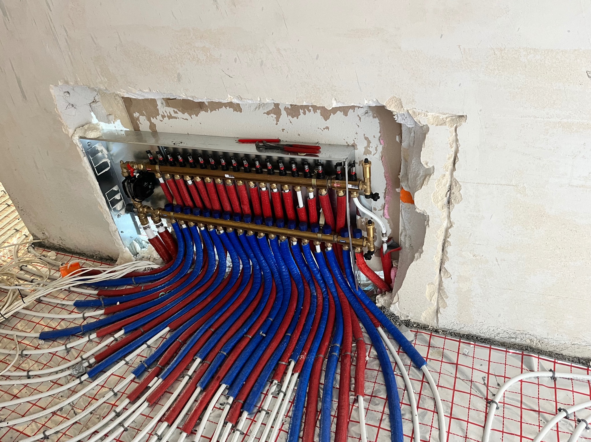 Rozdzielacz ogrzewania podłogowego z rurami w izolacji czerwonej i niebieskiej, zamontowany w surowej wnęce ściany, widoczne narzędzia na rozdzielaczu i mata grzewcza na podłodze.