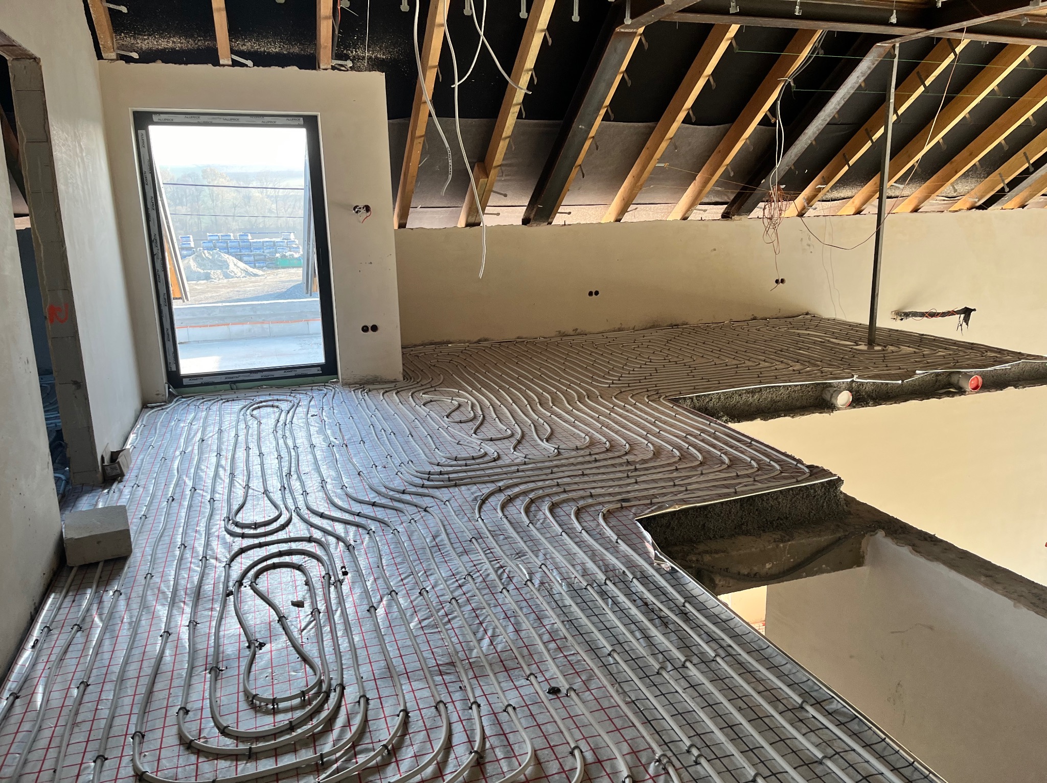 Niedokończona instalacja ogrzewania podłogowego z rurami ułożonymi w pętle na folii z siatką, widoczne surowe ściany, okno z widokiem na budowę i odsłonięta konstrukcja dachu.