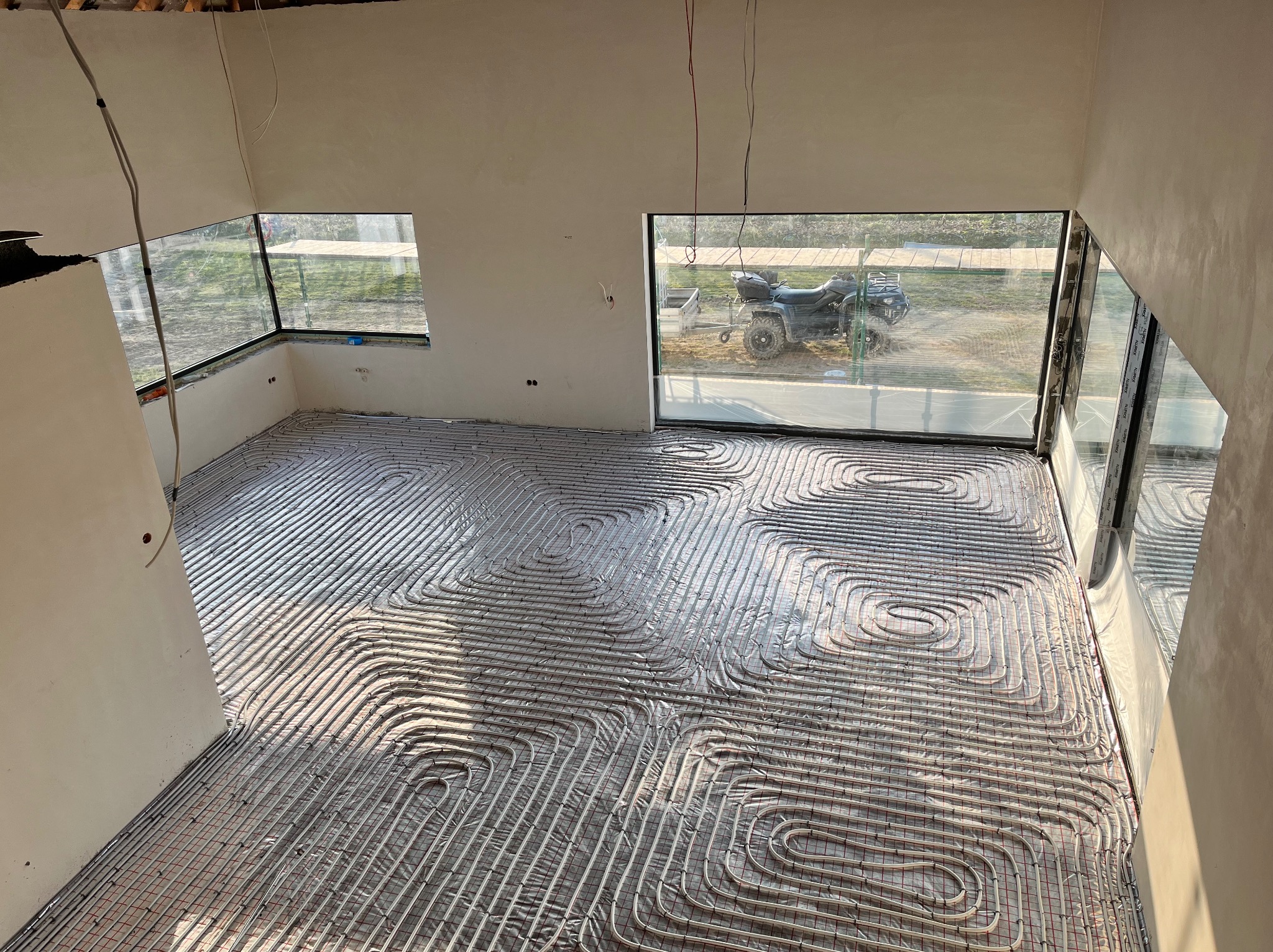 Rozłożona instalacja ogrzewania podłogowego w surowym pomieszczeniu, widok przez okno na quada z przyczepką.