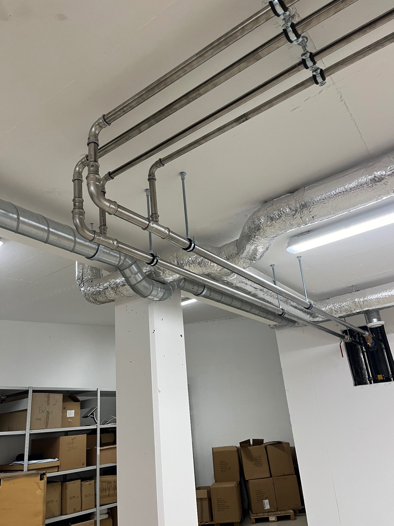 Rzędy rur ze stali nierdzewnej zamontowane pod sufitem, z widocznymi elementami mocującymi i izolacją termiczną, w tle regał z kartonami.