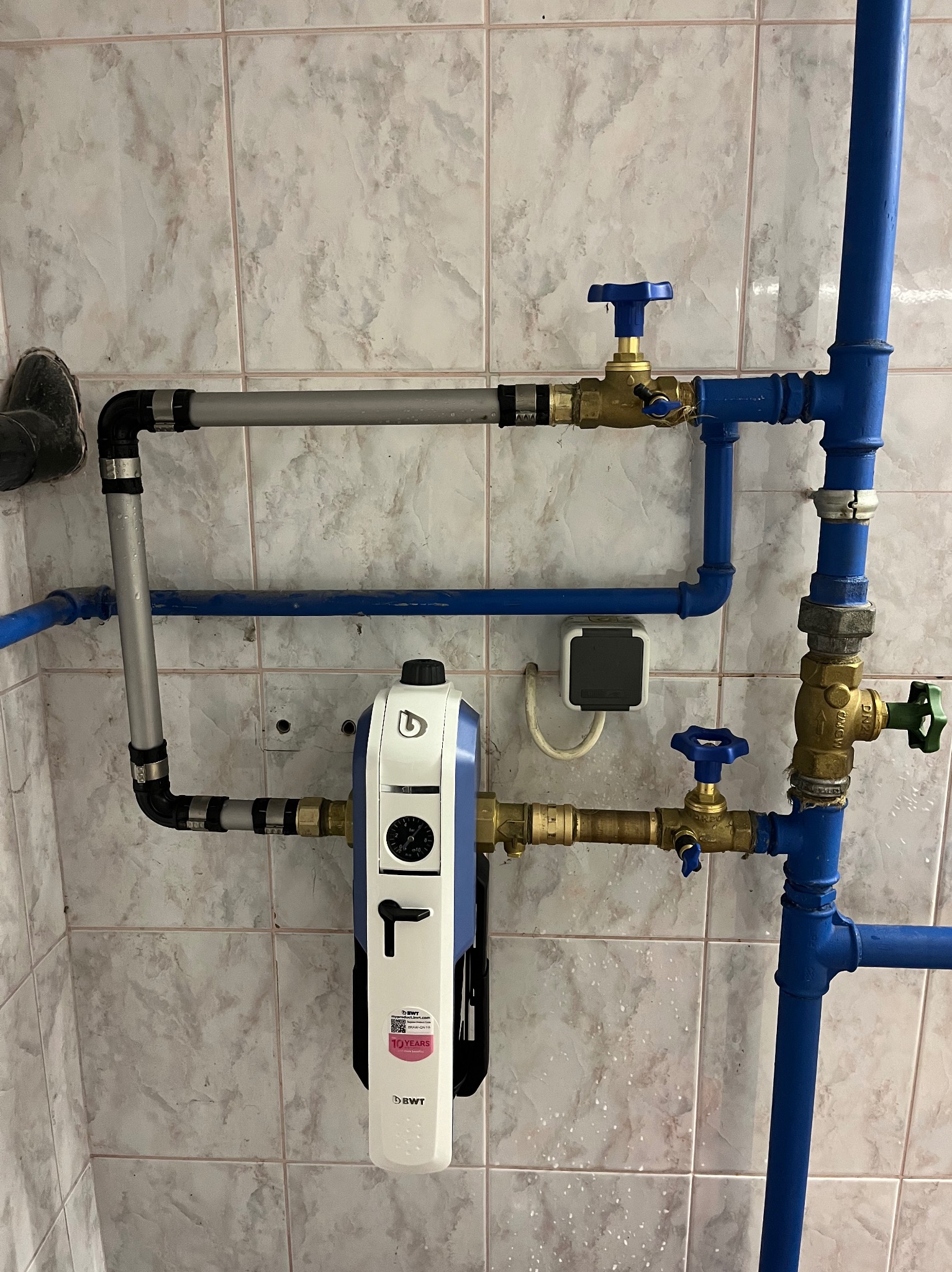 Szare i niebieskie rury wodne połączone z białym filtrem BWT z czarnym manometrem, zamontowane na ścianie z marmurkowych płytek, widoczne zawory.