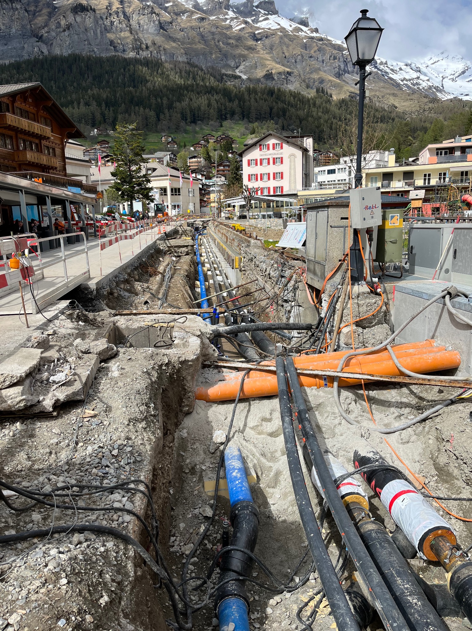 Otwarty wykop w miejskiej scenerii, ukazujący sieć rur i kabli w trakcie instalacji lub naprawy, z widocznymi elementami infrastruktury miejskiej w tle oraz górami w oddali.