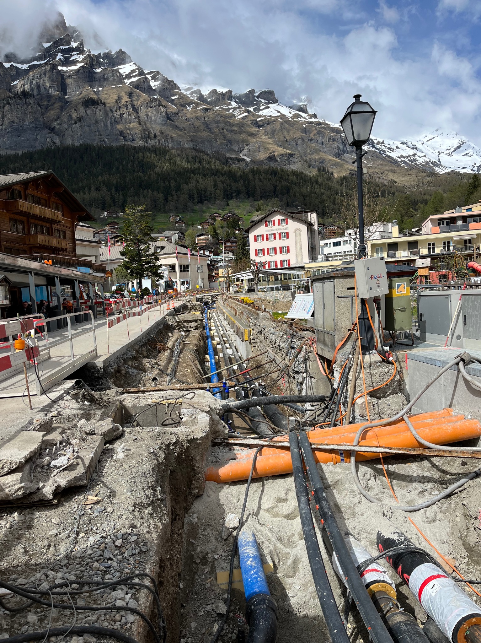 Wykonane wykopaliska pod sieć rur i kabli w malowniczej miejscowości górskiej, widoczne rury w kolorach niebieskim i pomarańczowym, kable oraz elementy betonowe.