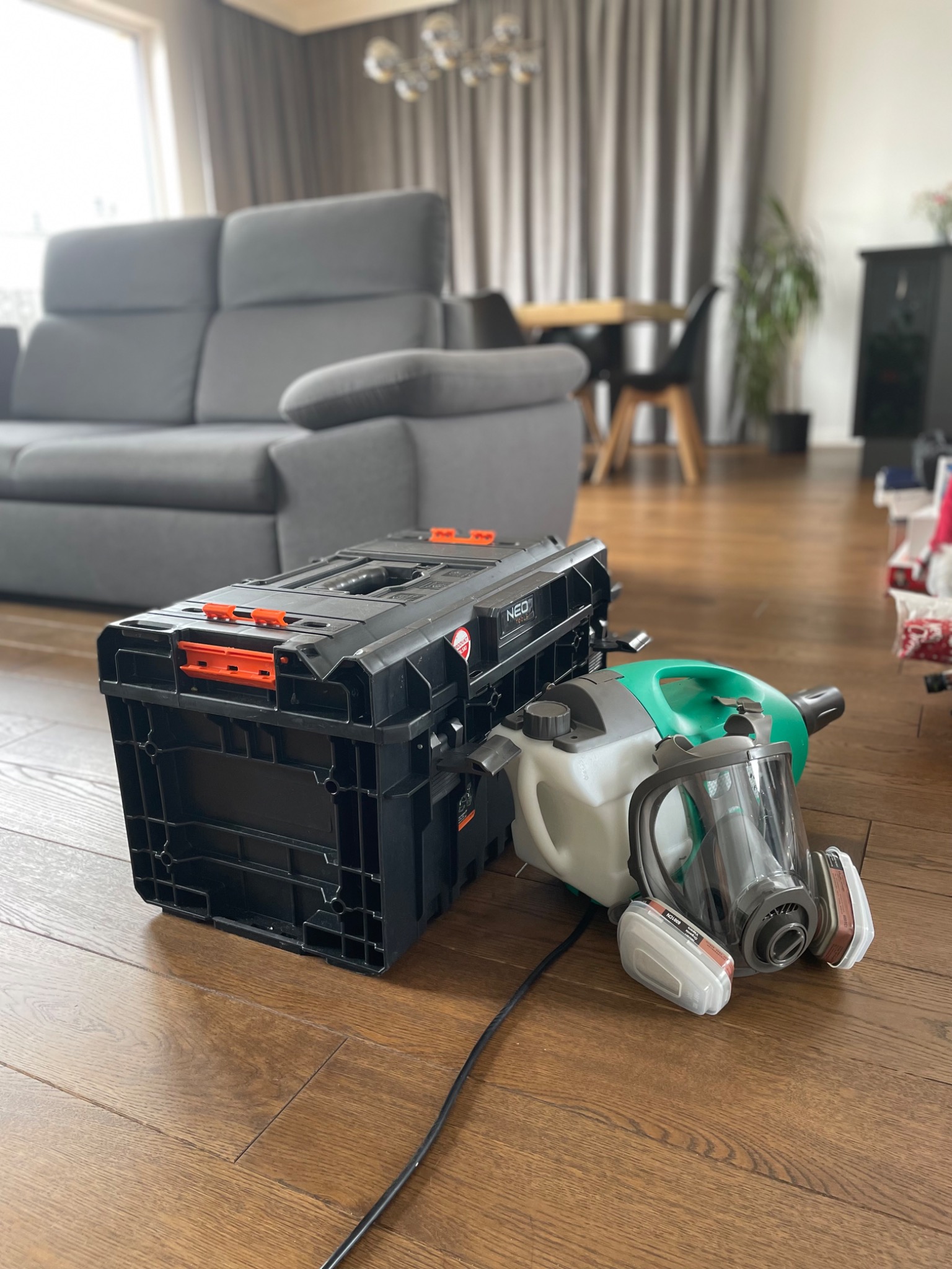 Sprzęt do zamgławiania ULV: maska ochronna, opryskiwacz i skrzynka narzędziowa NEO na drewnianej podłodze w salonie. Widoczny szary narożnik i stół z krzesłami.