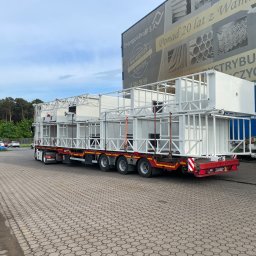 MST PROFI SPÓŁKA Z OGRANICZONĄ ODPOWIEDZIALNOŚCIĄ - Konstrukcja metalowa, biała, przestrzenna, modułowa, przewożona na naczepie ciężarówki na tle budynku z reklamą firmy Drozapol-Profil S.A.