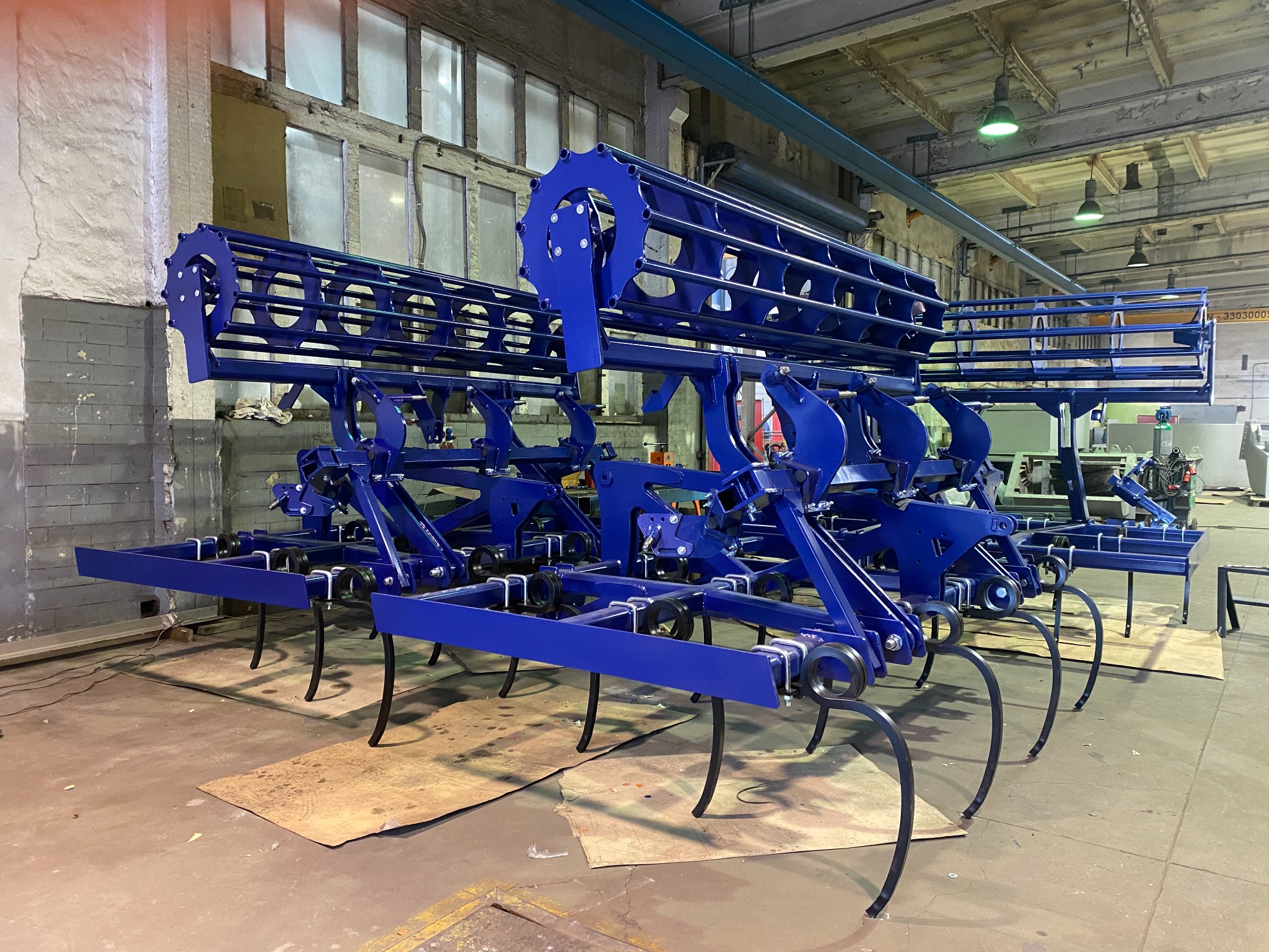 Niebieski agregat uprawowy z wałem strunowym w hali produkcyjnej, widoczne elementy metalowe i spawane połączenia. Sprzęt rolniczy, produkcja maszyn.