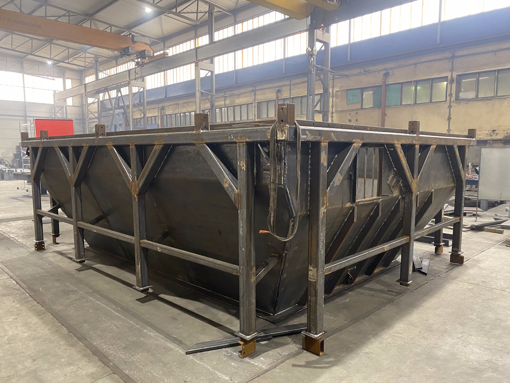 Konstrukcja stalowa w hali produkcyjnej, spawana z profili i blach, podwieszona na suwnicy. Widoczne ślady spawania i obróbki metalu. Surowy, industrialny charakter.