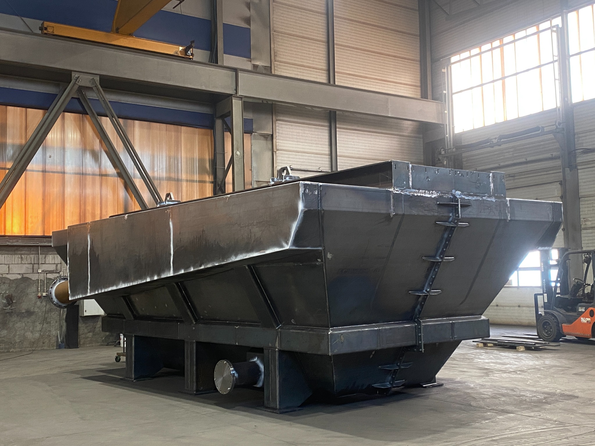 Duży, surowy, metalowy zbiornik przemysłowy z widocznymi spawami, umieszczony w przestronnej hali produkcyjnej z oknami i wózkiem widłowym w tle.