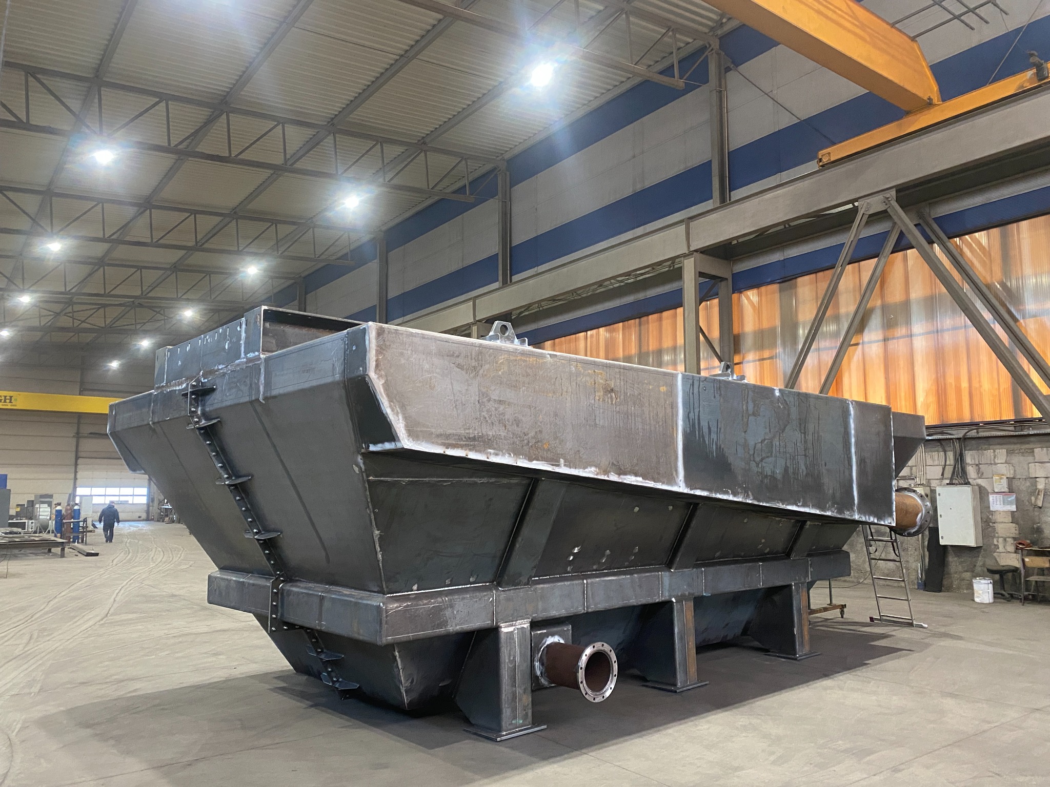 Surowa, stalowa konstrukcja zbiornika z rurami, spawana w hali produkcyjnej. Widoczne ślady spawania, drabinka i fragment suwnicy. W tle pracownik i elementy warsztatu.