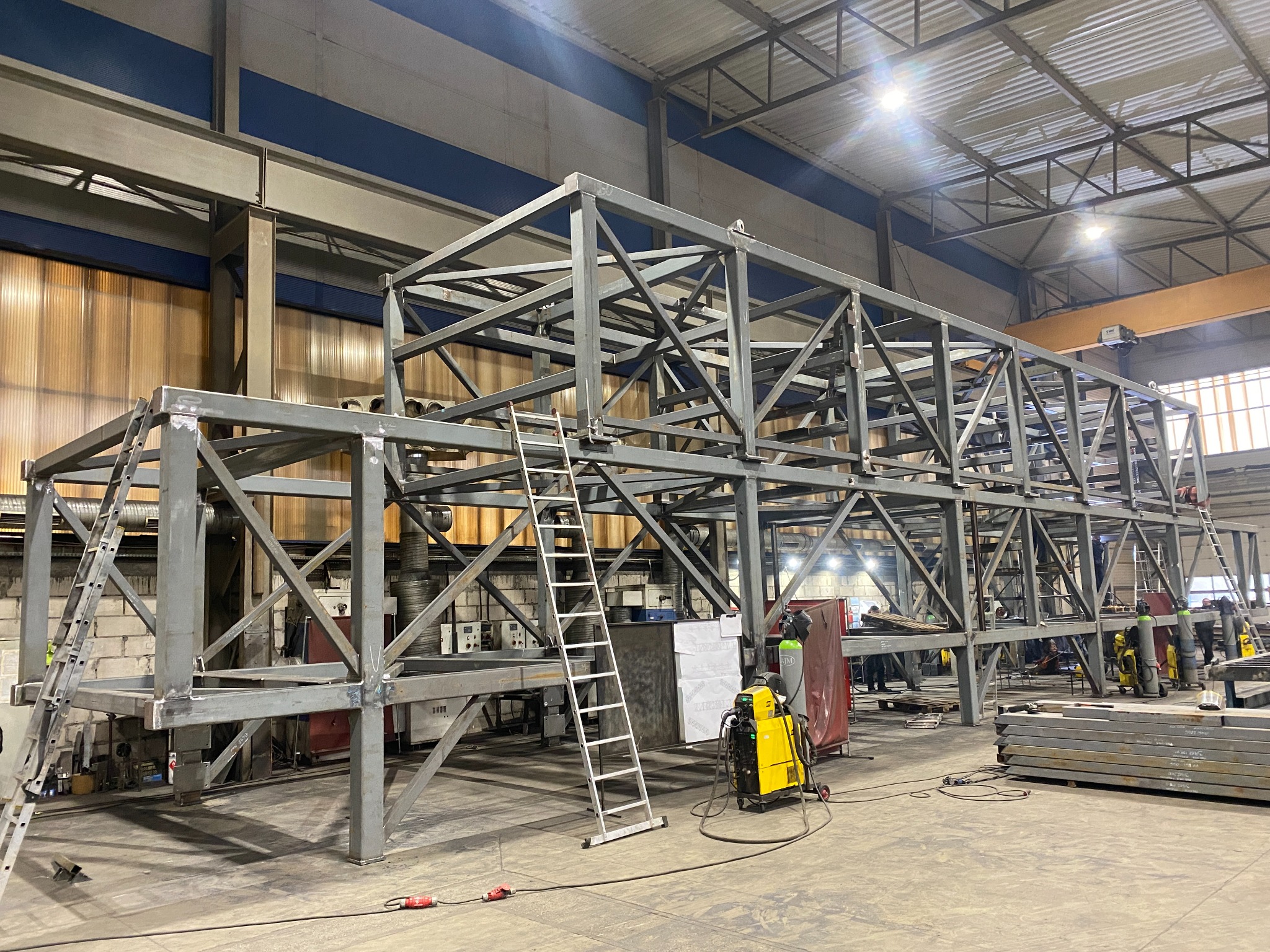 Szkielet metalowej konstrukcji, spawana rama z drabinką w hali produkcyjnej. Widoczne narzędzia spawalnicze i elementy konstrukcyjne.