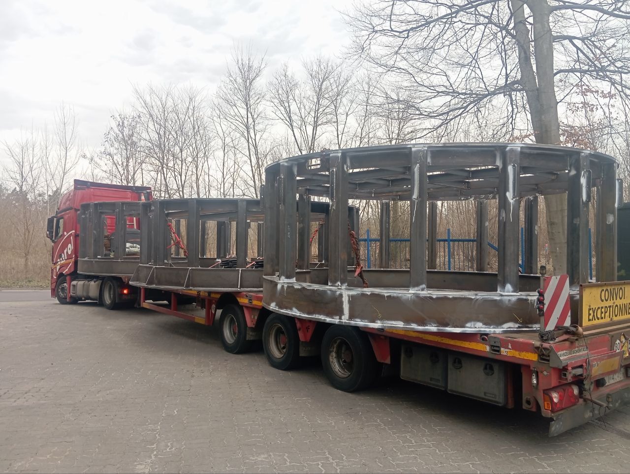 Transport dwóch metalowych konstrukcji o okrągłym kształcie na naczepie ciężarówki. Widoczne spawy i surowy metal. Tablica ostrzegawcza na tyle naczepy.