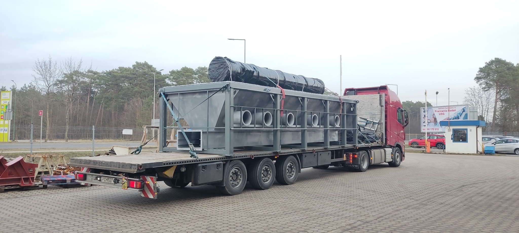 Konstrukcja metalowa z rurami na naczepie ciężarówki, zabezpieczona pasami transportowymi. W tle las i budynki. Elementy konstrukcyjne w kolorze szarym.