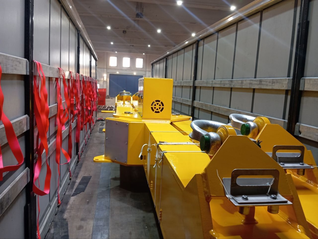 Żółta konstrukcja metalowa z uchwytami i szeklami, zabezpieczona czerwonymi pasami wewnątrz hali produkcyjnej. Widoczne oświetlenie sufitowe i metalowe ściany.