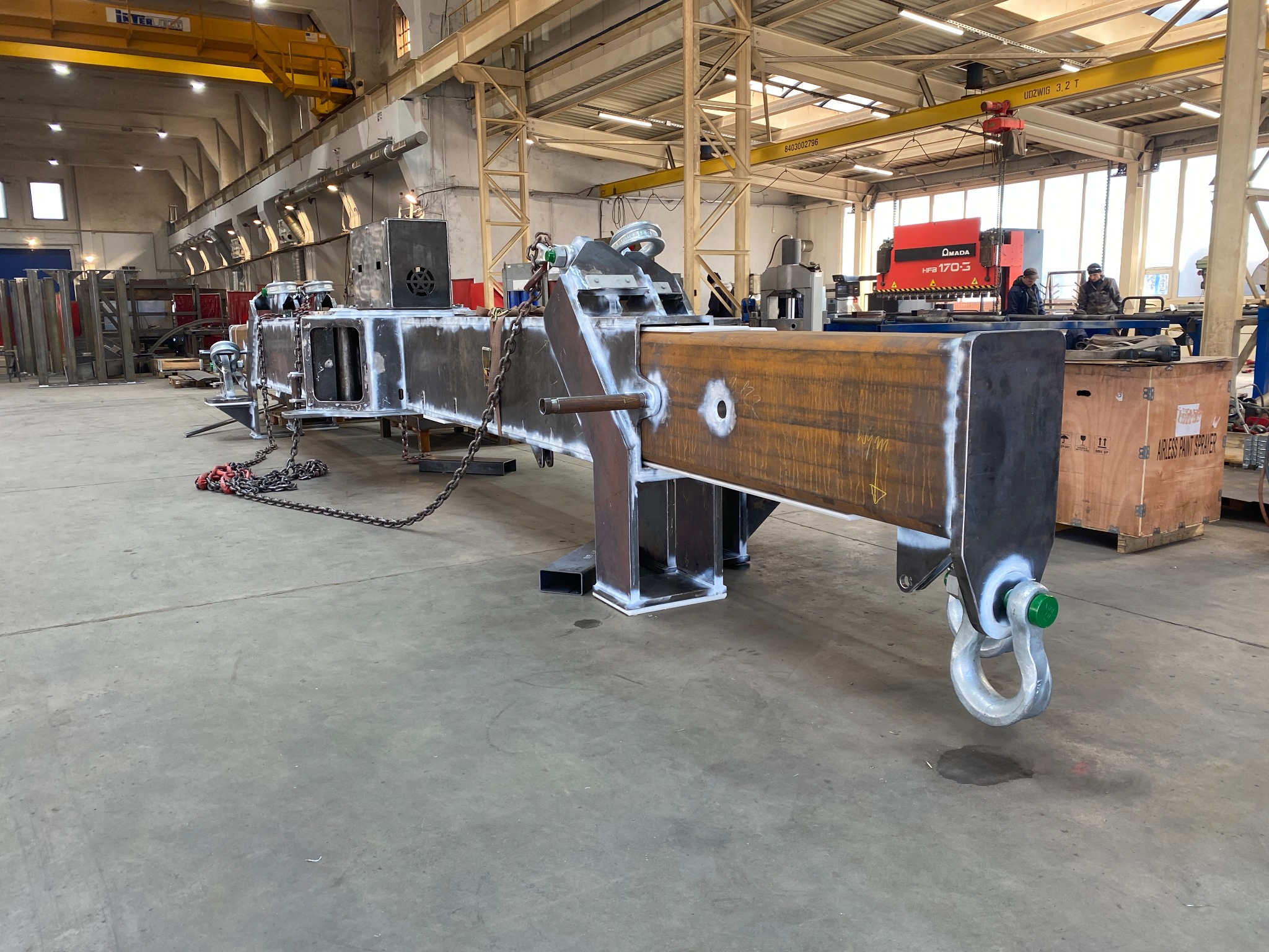 Surowy, spawany element konstrukcyjny z metalu z hakiem, łańcuchami i otworami, w hali produkcyjnej z suwnicami i maszynami w tle. Widoczne ślady spawania.